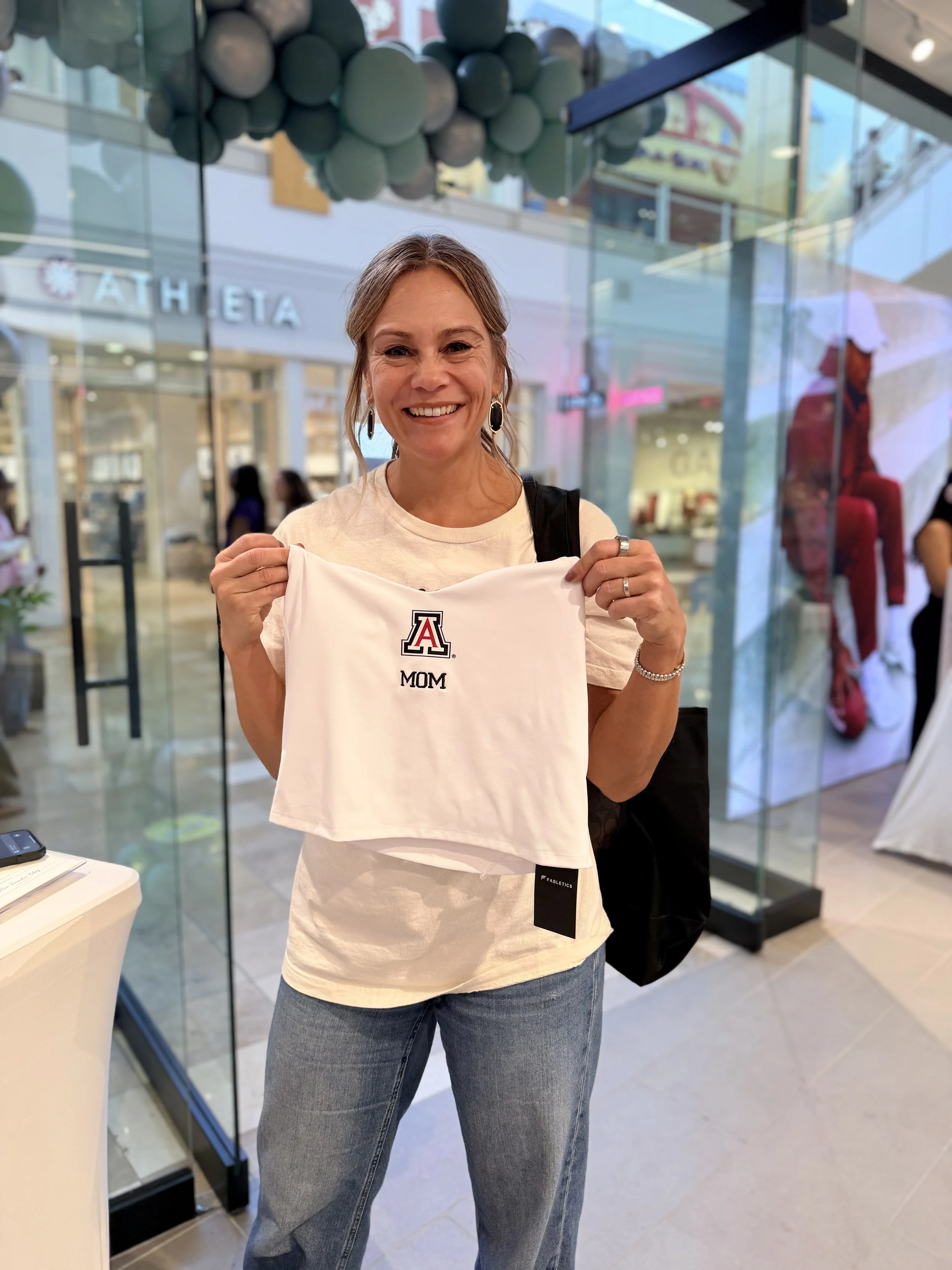 A smiling woman holding a white cloth with a University of Arizona logo and the word "Mom" in front of a glass storefront with decorative green balls hanging from the ceiling inside a shopping mall.