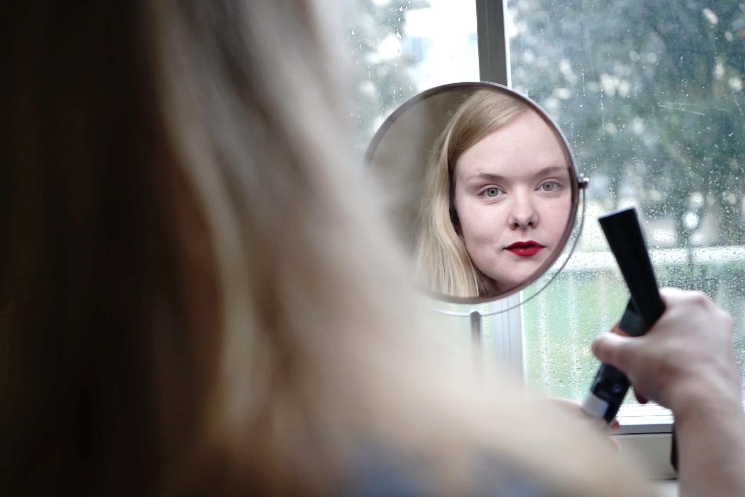 A woman looking into a small round mirror as she applies red lipstick, with a window showing raindrops in the background.