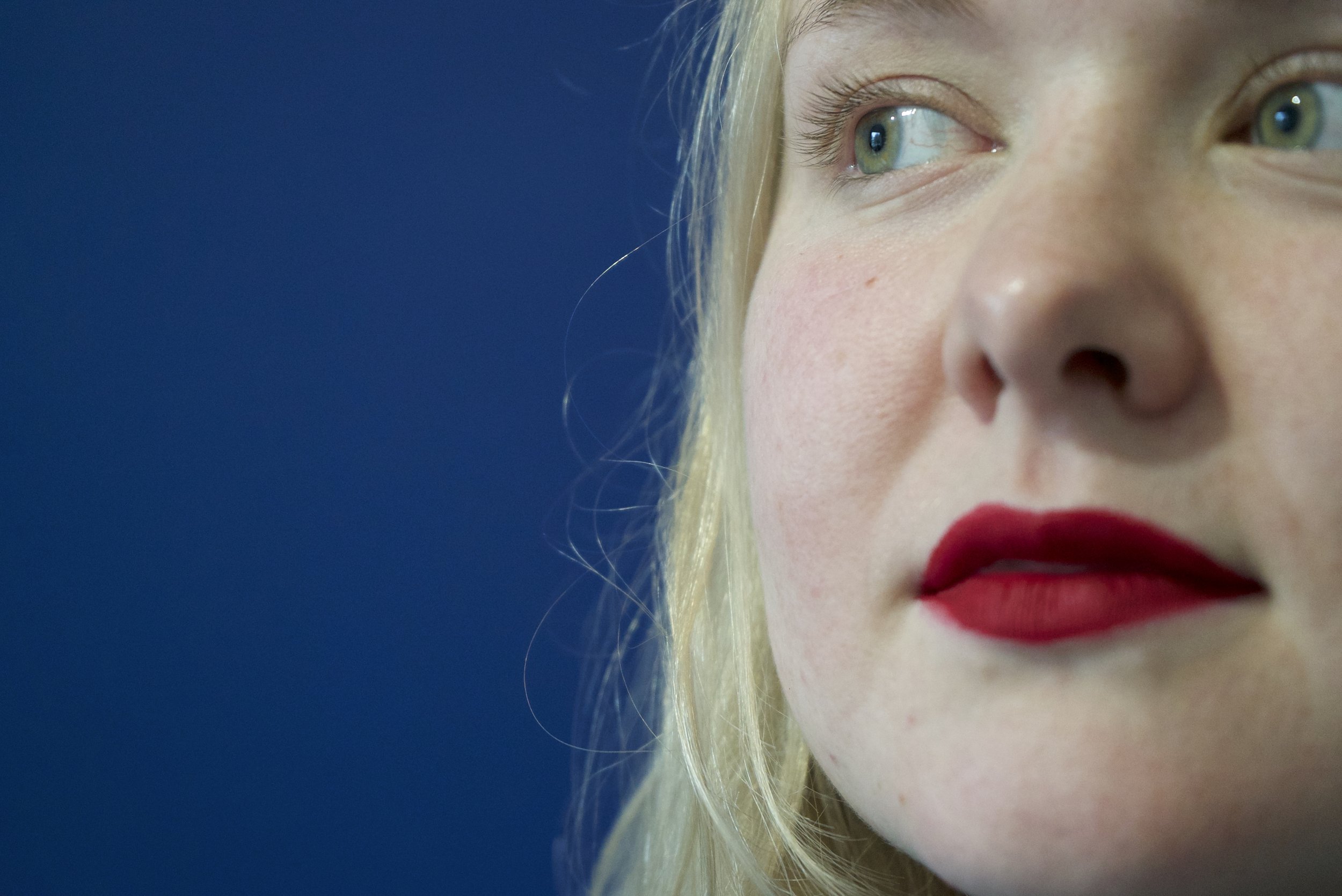 A close-up photograph of a woman with blonde hair, red lipstick, and green eyes, against a dark blue background.