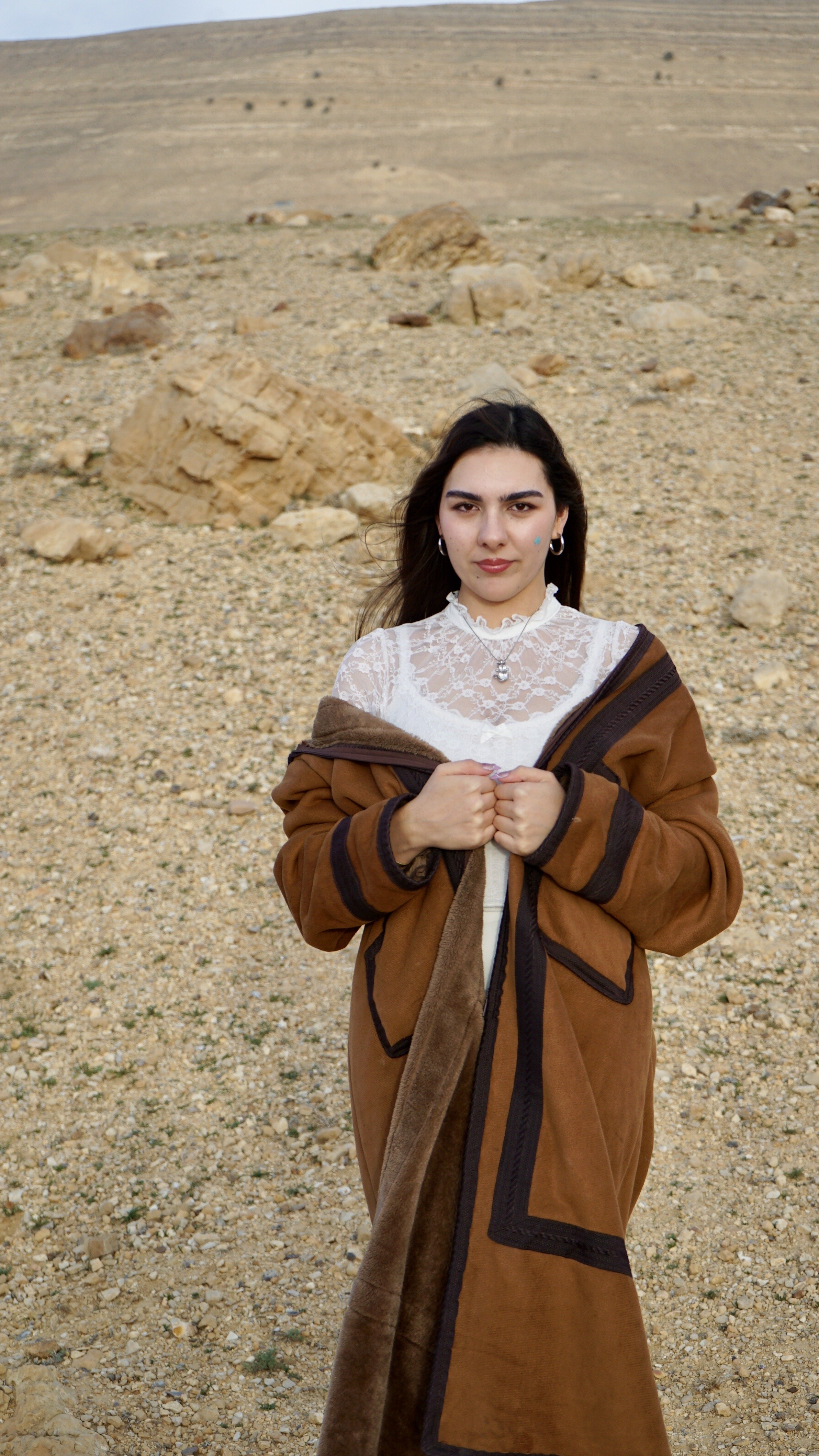 A young woman standing outdoors in a rocky, desert-like landscape with layered hills in the background, wearing a white lace top and a brown coat, with her hands gripping the coat closed, and looking directly at the camera.
