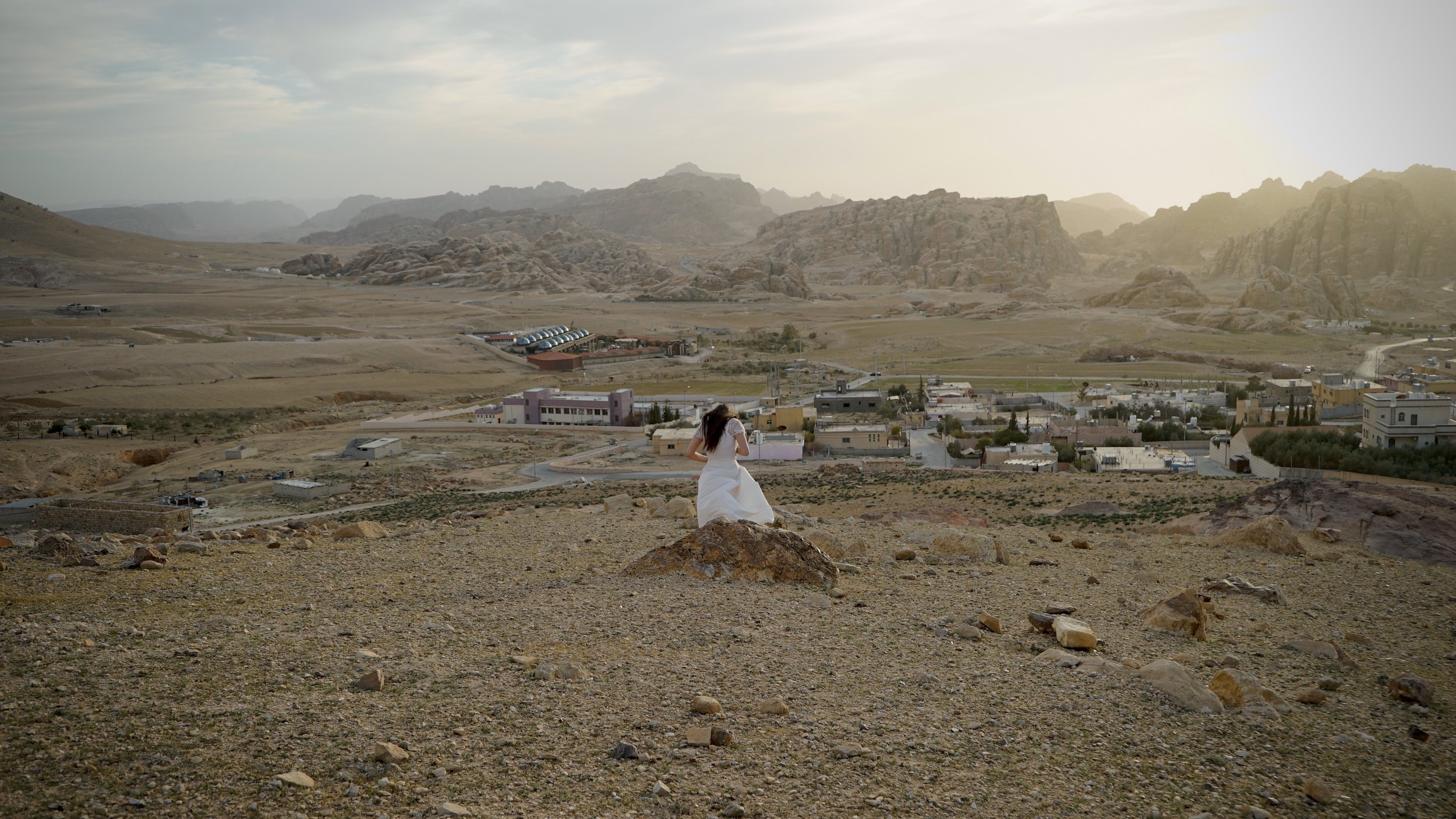 A woman in a white dress standing on a rock in a desert landscape overlooking a small town and mountain ranges in the distance during sunset or sunrise.