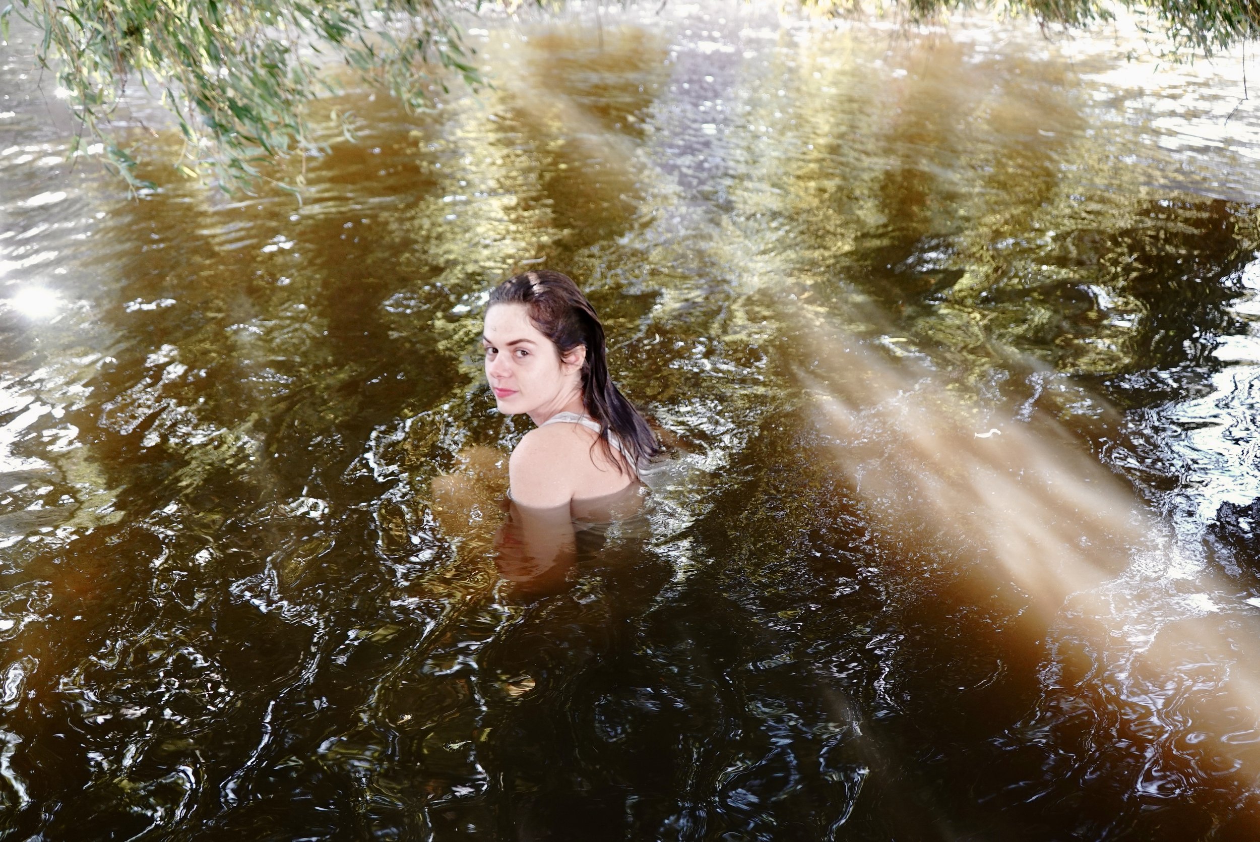 A young woman with dark hair in a pond surrounded by greenery, looking at the camera.