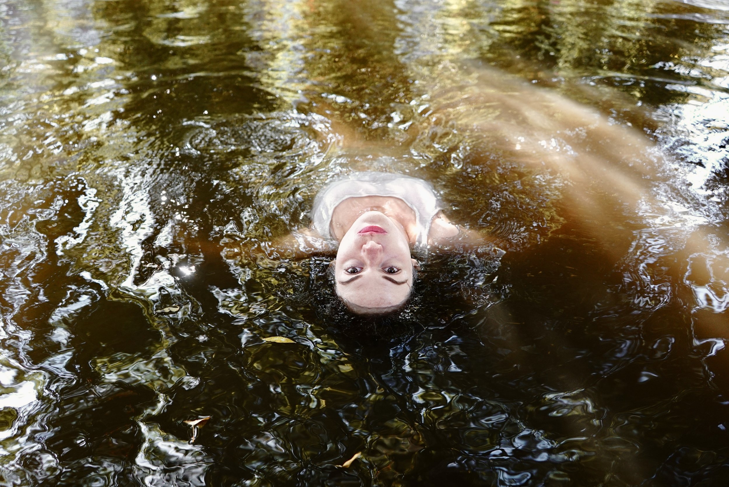 A woman with blonde hair floating on her back in a dark river, looking up at the camera with her hair spread out in the water.