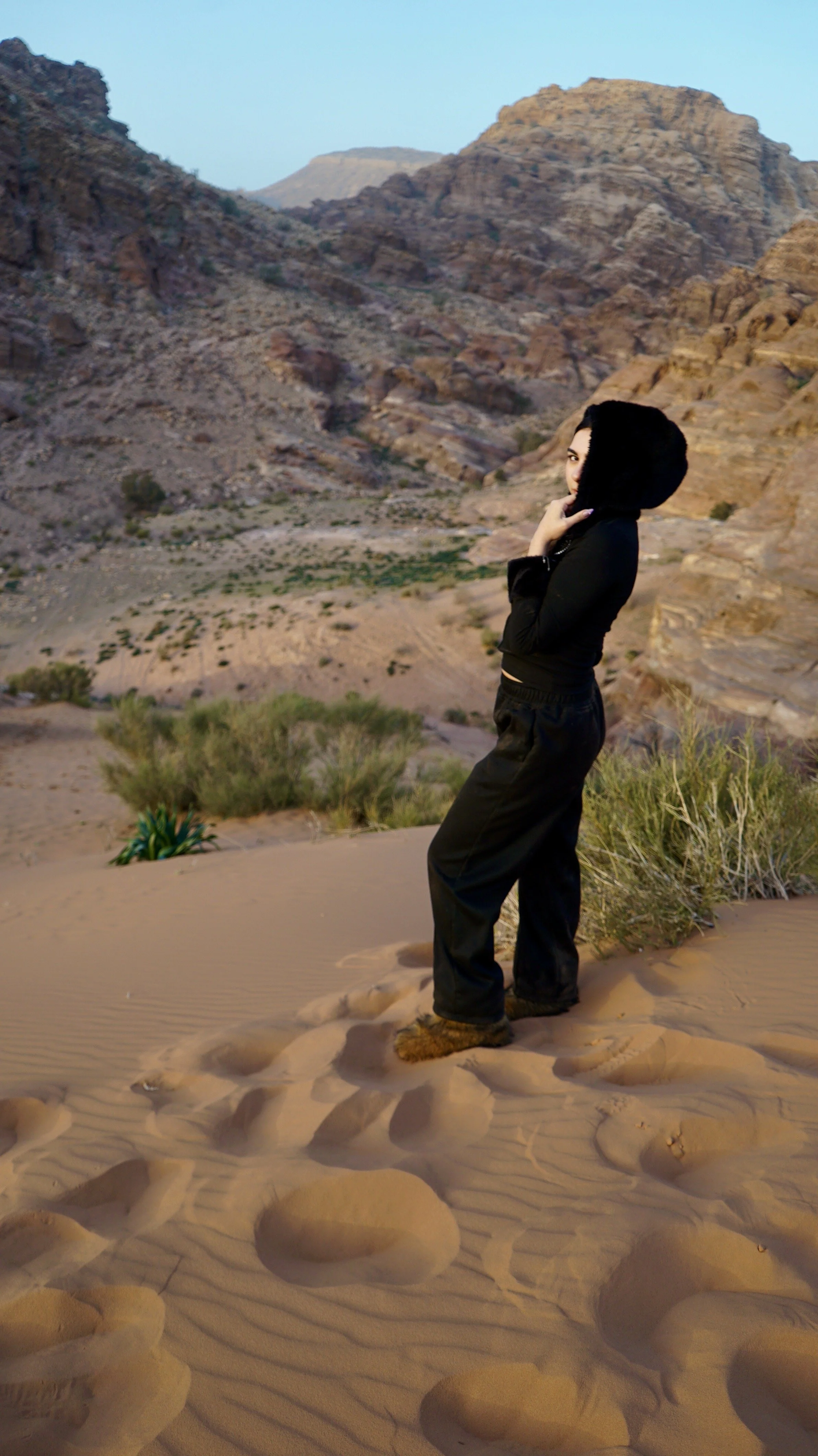 A woman dressed in black standing in a desert surrounded by sand dunes, desert plants, and rocky mountains under a clear sky.