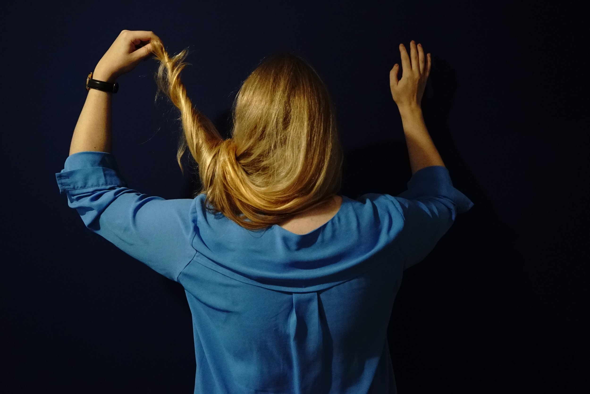 Back view of a woman with long red hair, wearing a blue shirt, standing against a dark background, holding a strand of her hair.