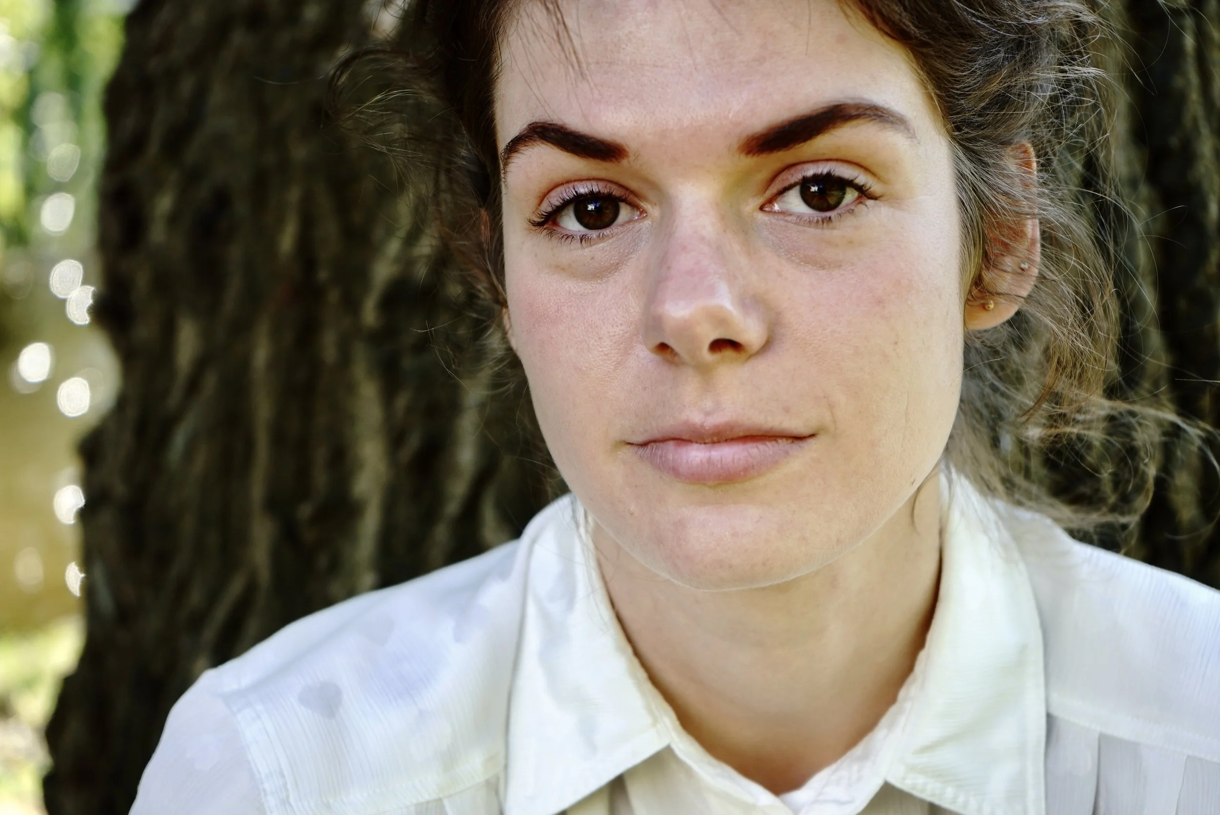 Close-up of a young woman with dark hair and brown eyes, wearing a white shirt, outdoors near a tree.