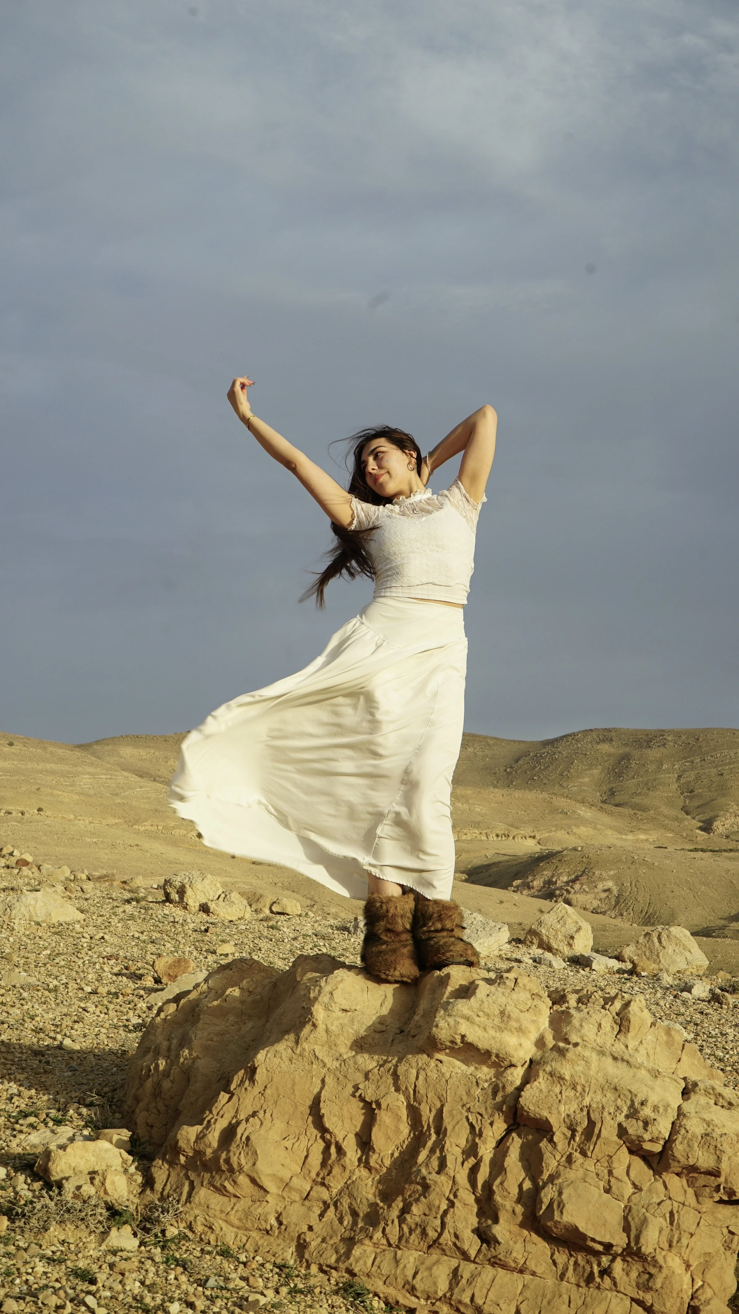 A woman in a white dress and fur boots standing on a large rock in a desert landscape with a cloudy sky.