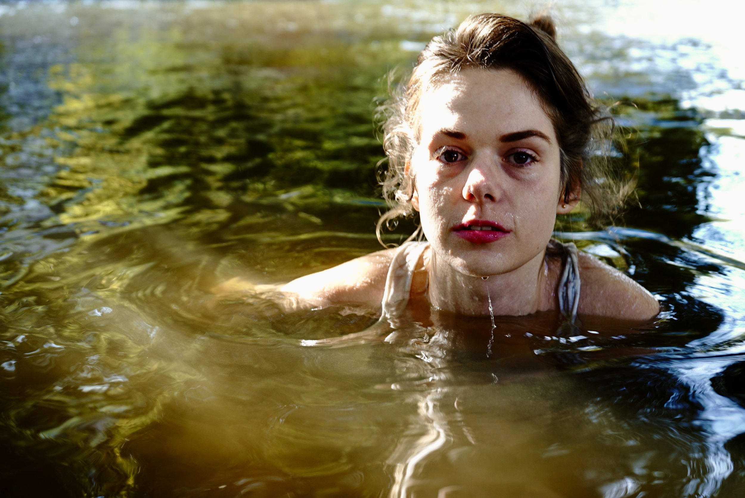 A young woman with wet hair and light skin submerged in a body of water, looking directly at the camera with a serious expression, during daylight.