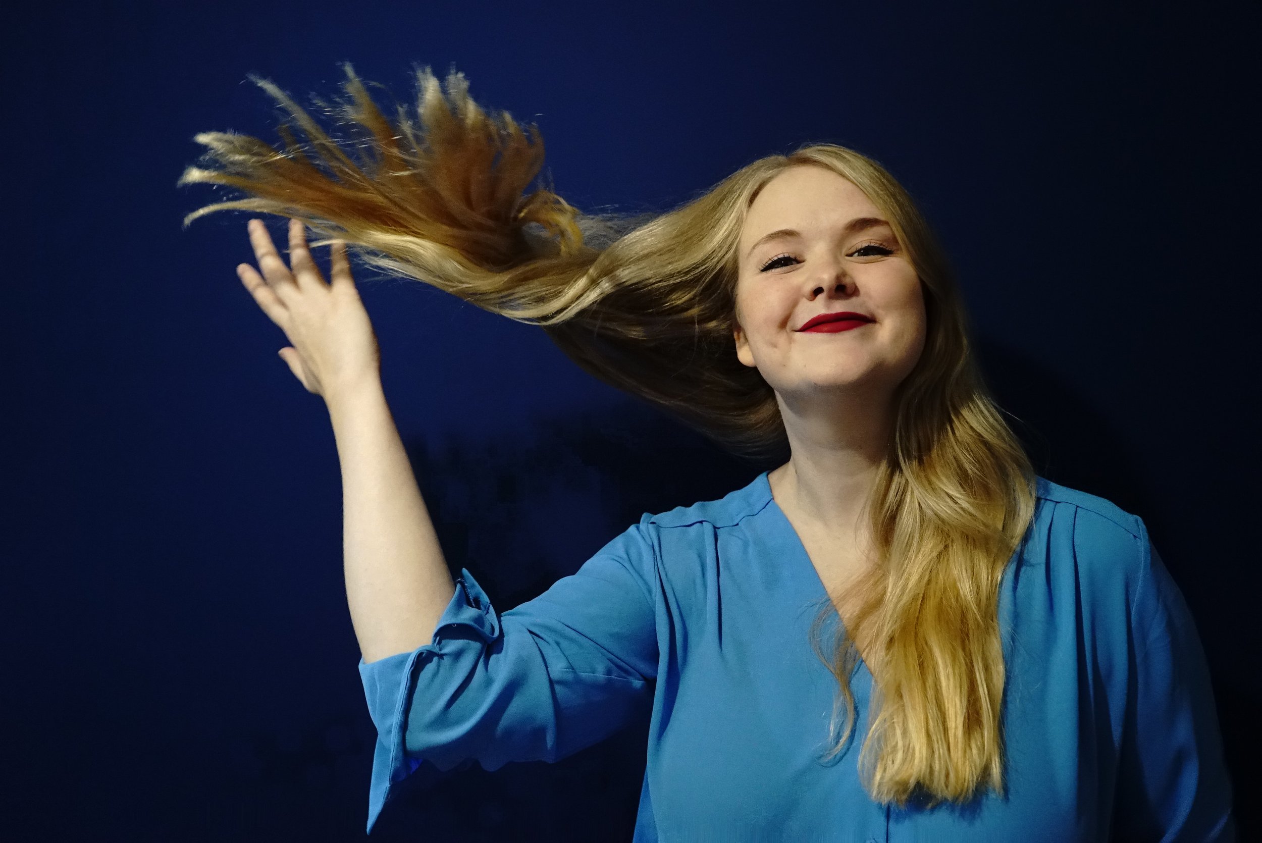 A young woman with long blonde hair, wearing a blue shirt, smiling and tossing her hair against a dark background.