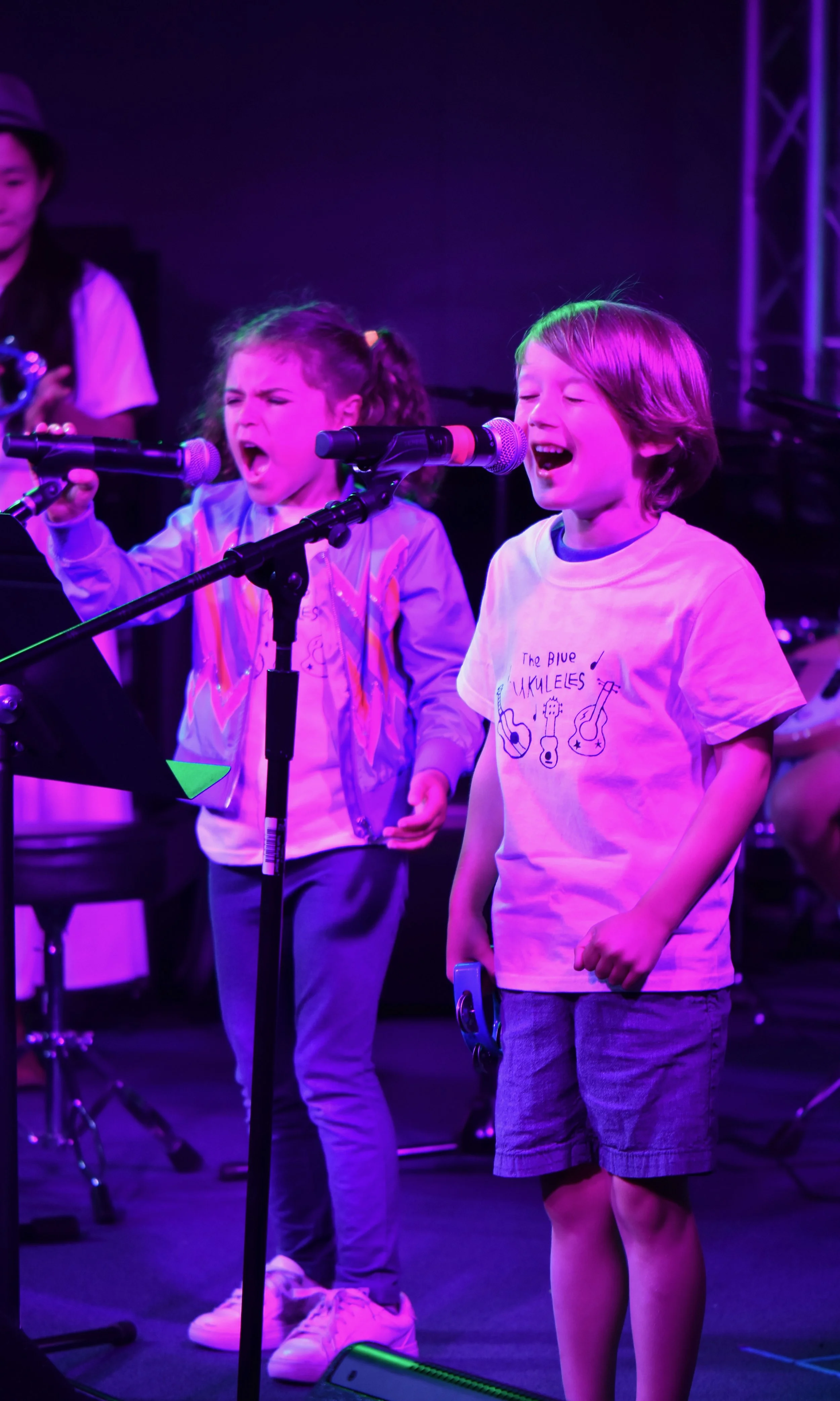 Two children singing into microphones on stage, with a girl wearing a T-shirt reading "The Blue Ukuleles" and a boy with long hair, in a colorful, vibrant setting.