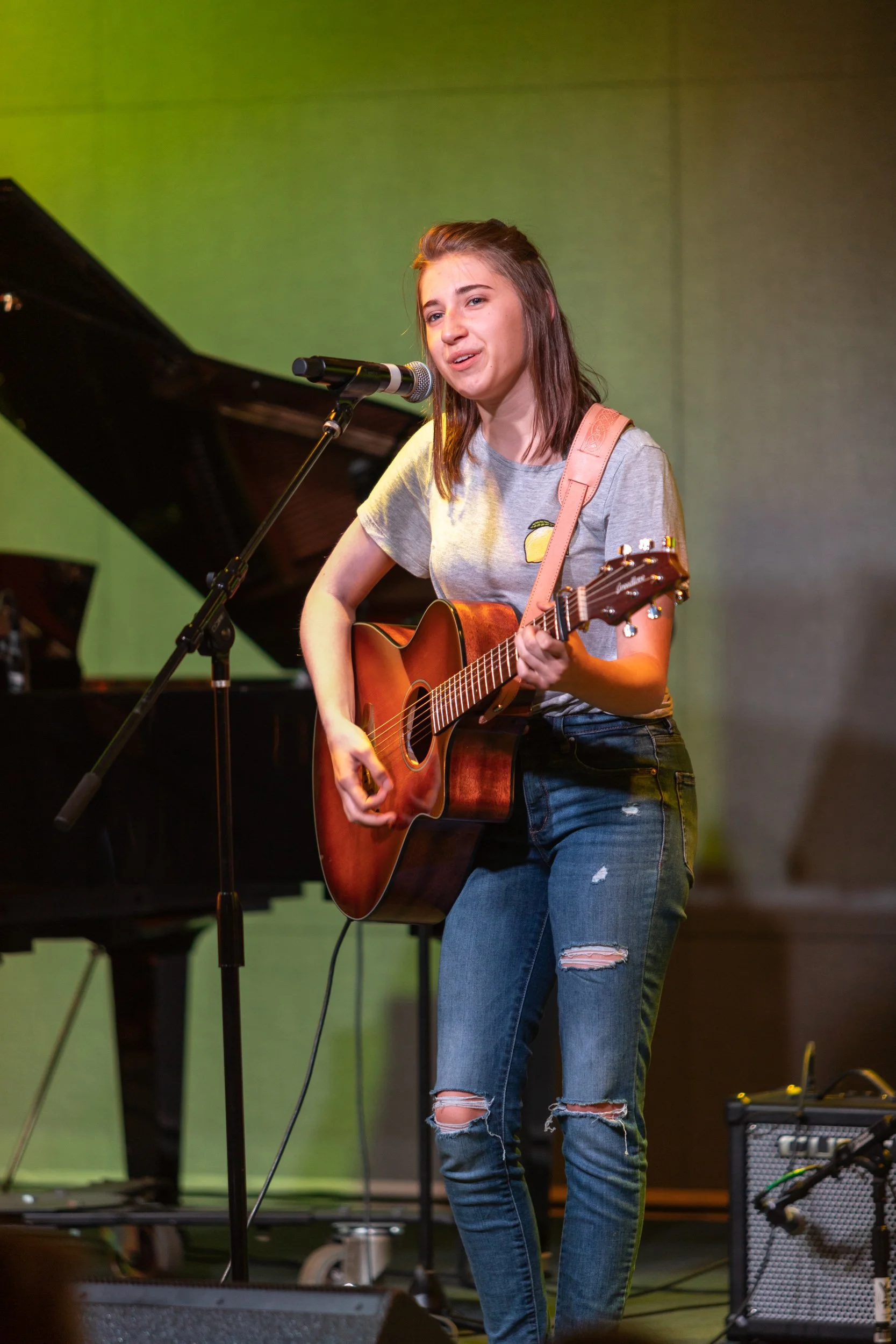 Young woman playing an acoustic guitar and singing into a microphone on stage, with a grand piano in the background.