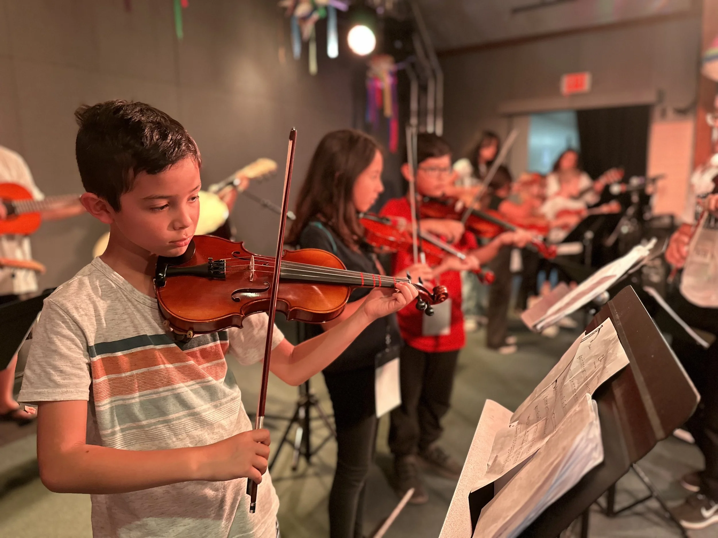 A group of children playing violins in a music class or rehearsal, with music sheets on stands in front of them.