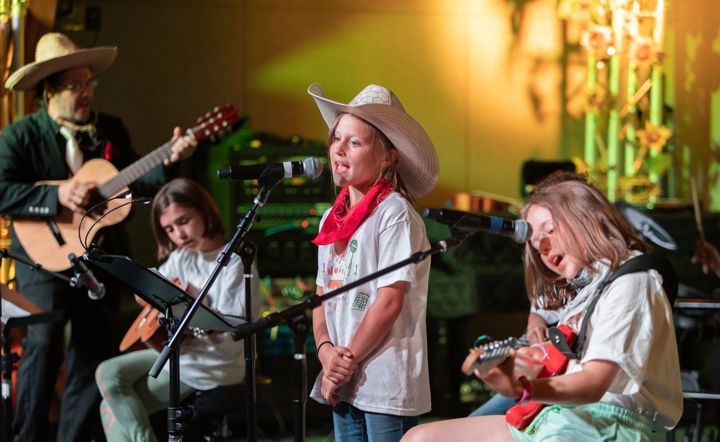 Children performing with musical instruments on stage, singing into microphones, wearing casual clothes and hats, with a man playing a guitar in the background, colorful stage lighting, and musical equipment.