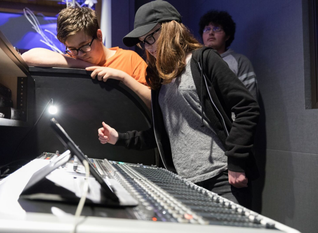 Three young people, two boys and a girl, are in a music or recording studio. The girl is at a sound mixing console, adjusting controls, wearing a black hoodie, gray shirt, glasses, and a black cap. One boy is leaning over a soundproof divider, and the other boy stands behind them observing.