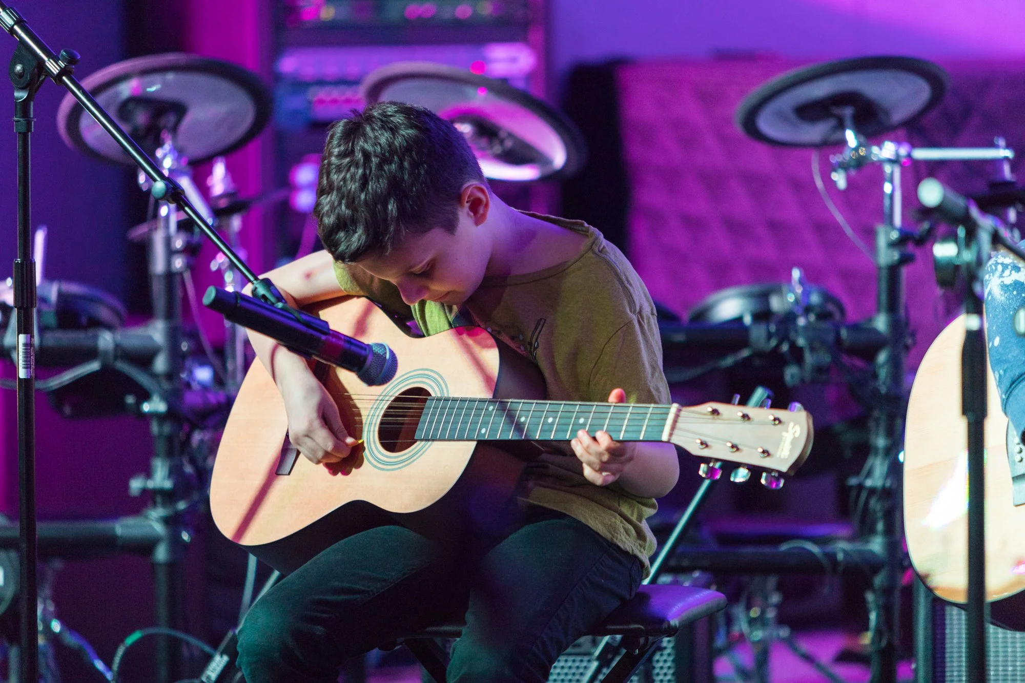 A young person sitting on a chair, playing an acoustic guitar on a stage with purple lighting and musical equipment in the background.