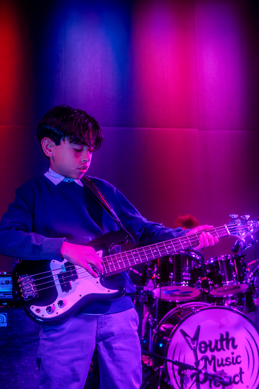 A young boy playing a bass guitar during a music performance, with a drum set in the background branded 'Youth Music Project', under colorful stage lighting.