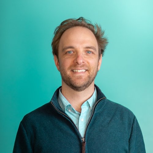 Portrait of a smiling man with light brown hair, beard, wearing a light blue collared shirt and a dark zip-up sweater against a teal background.