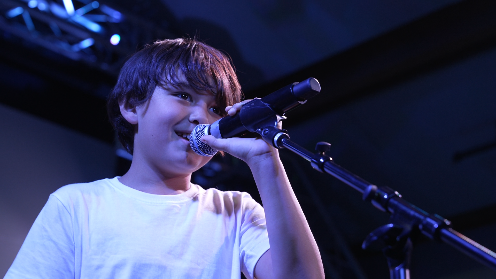 Young boy singing into a microphone on stage, wearing a white t-shirt, with stage lighting in the background.