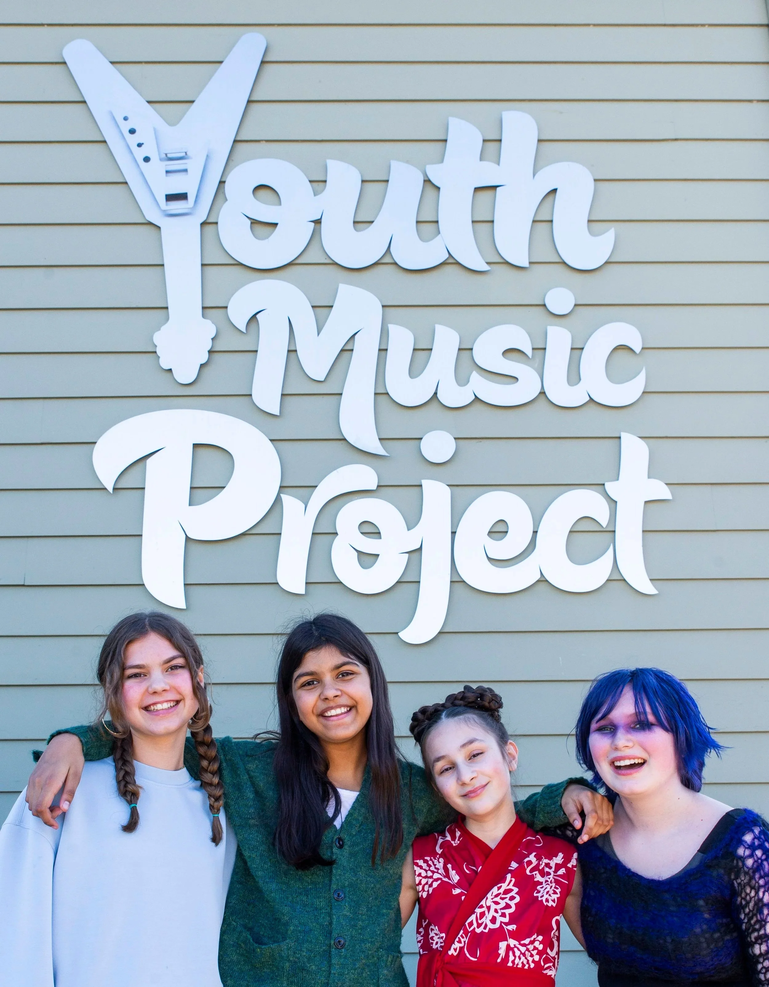 Group of diverse young girls smiling, standing arm in arm in front of a wall with a sign that reads 'Youth Music' and features a large peace sign.
