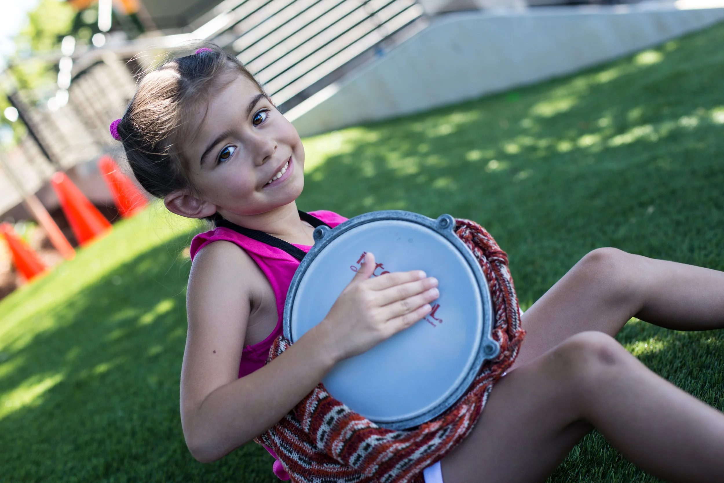 A young girl with brown hair and a pink shirt sitting on grass, smiling at the camera, holding a white drum.