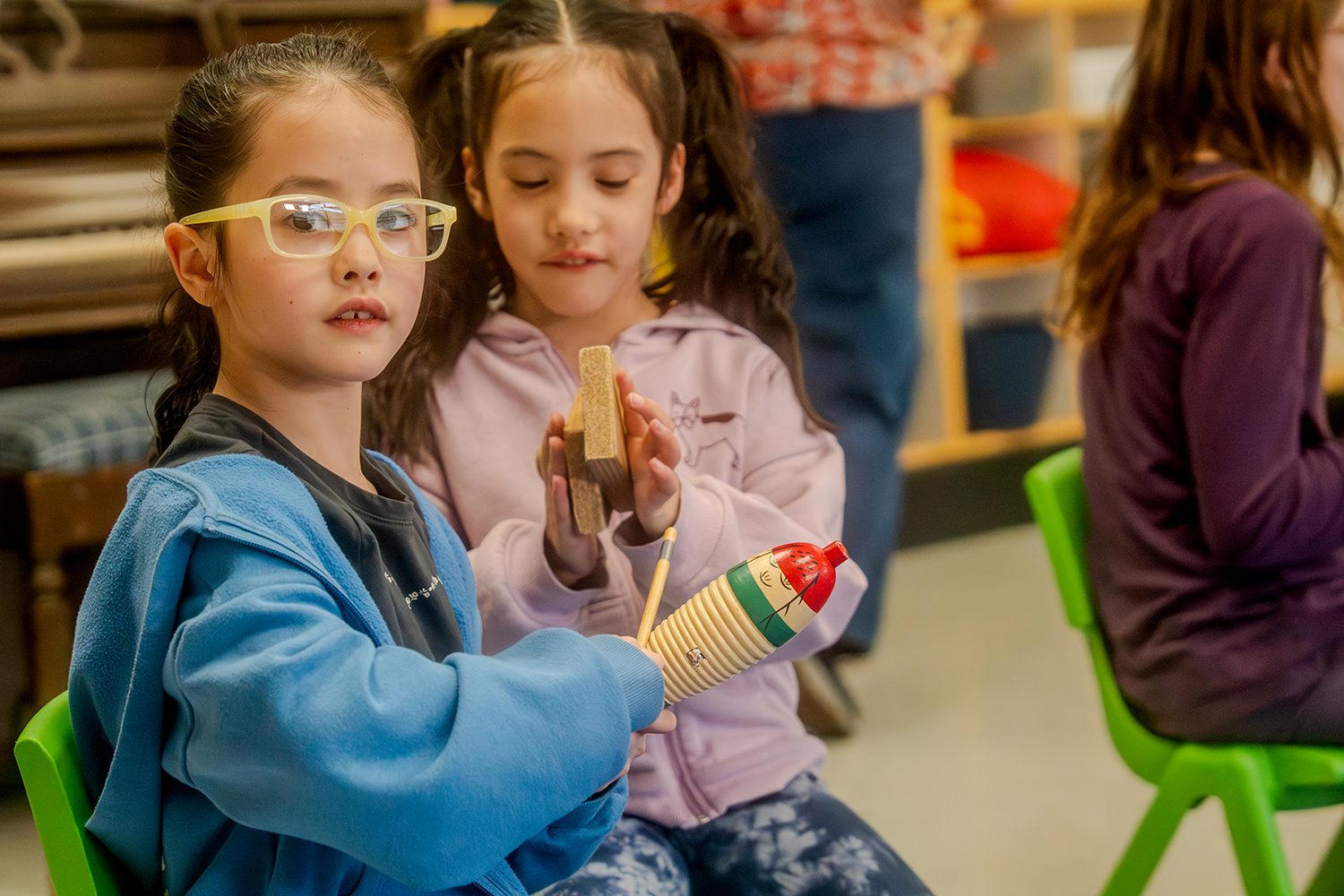 Two young girls sitting on green chairs in a classroom, one wearing glasses and a blue hoodie holding a small musical instrument, the other in a pink hoodie holding wooden blocks.