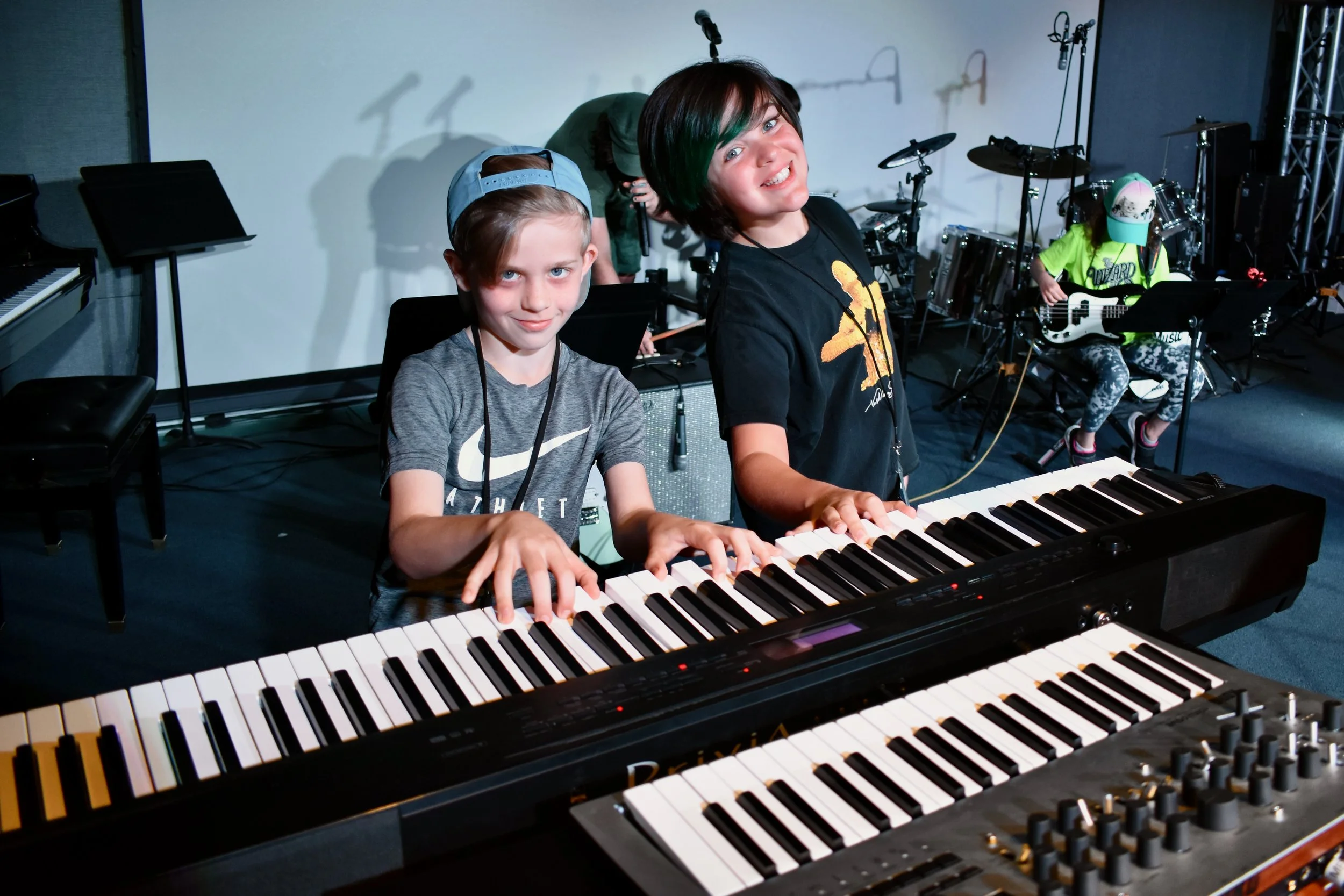 Two boys playing keyboard instruments on stage with musical equipment in the background.