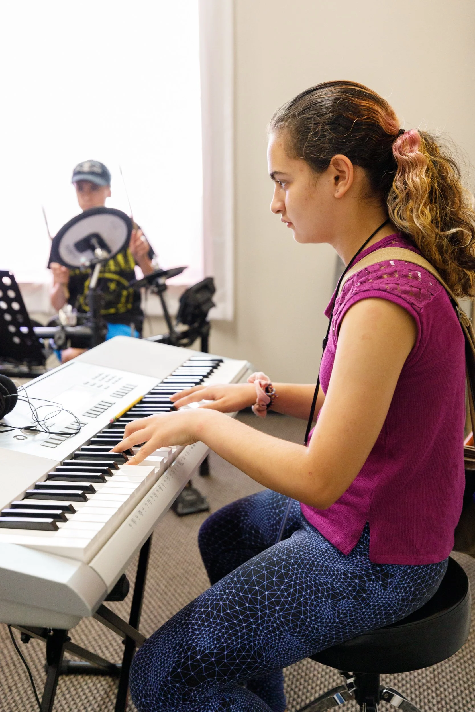 A young woman with curly hair playing a white electronic keyboard in a music rehearsal room. She is wearing a pink top with cut-out details and geometric-patterned pants, focusing on her playing. In the background, a male musician with a cap and a black and yellow shirt is adjusting equipment.