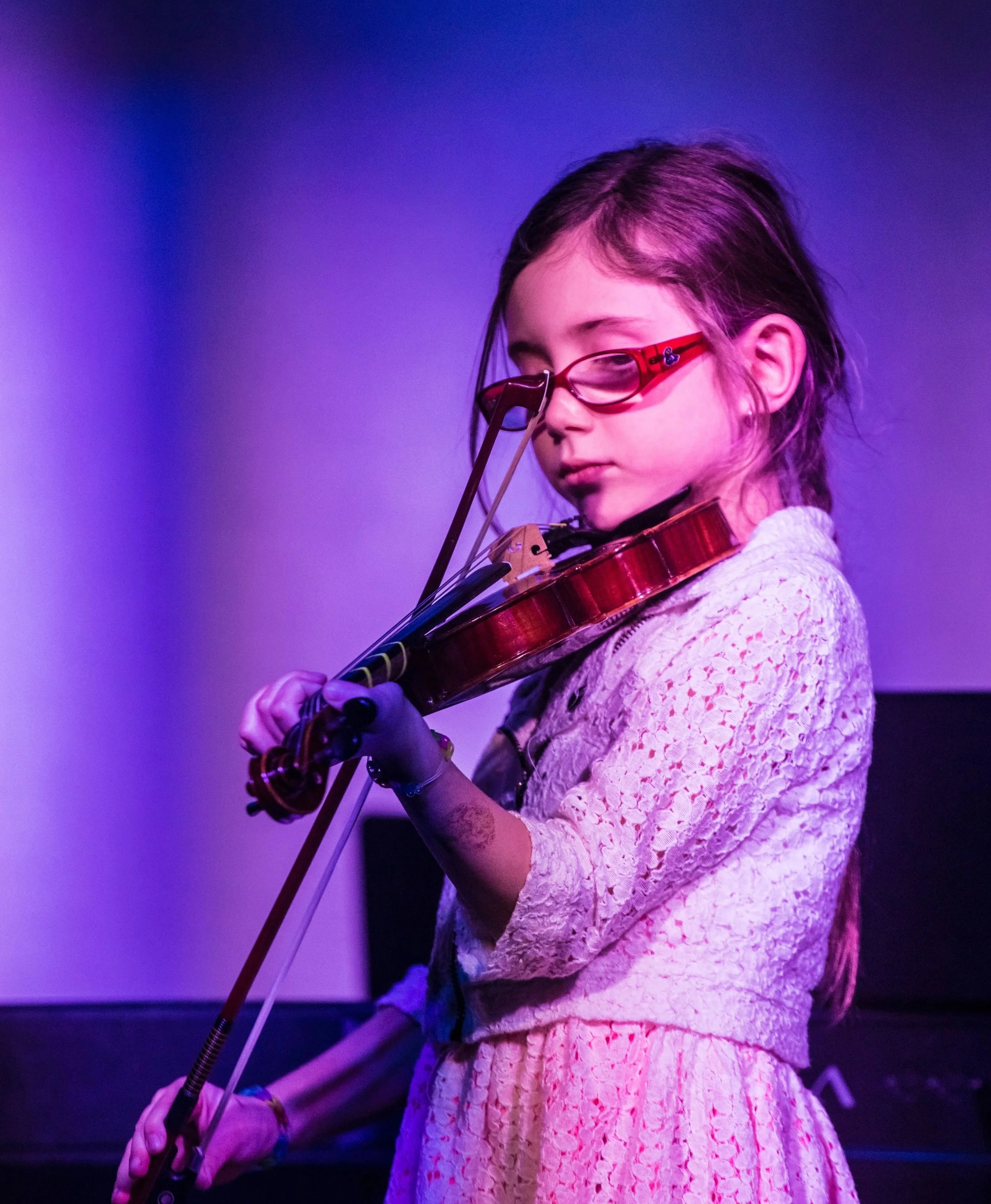 A young girl with long hair and glasses playing a violin against a purple-lit background.
