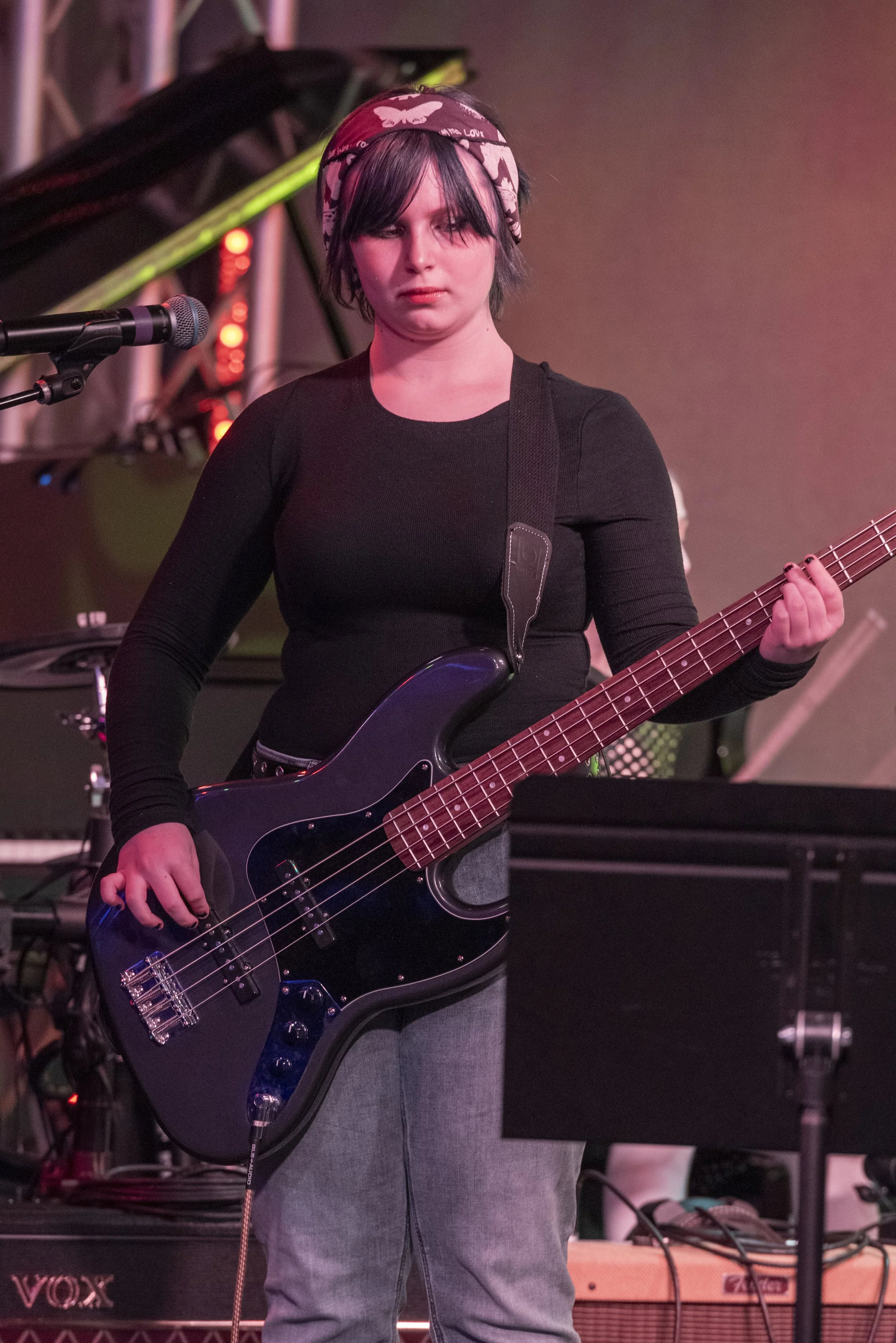 A young woman with short dark hair wearing a pink butterfly-patterned headband, playing a black electric bass guitar on stage with a microphone in front of her. She is dressed in a black long-sleeved shirt and gray pants, with stage equipment and a keyboard visible in the background.