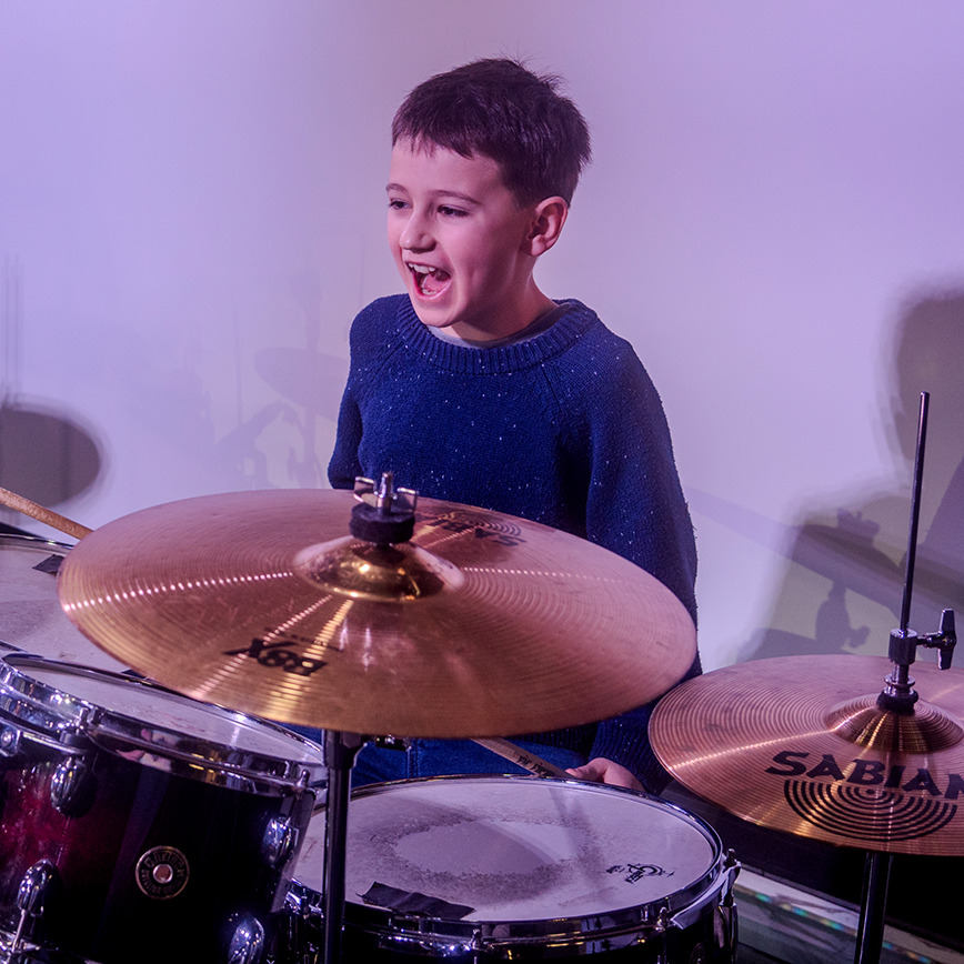 A young boy playing a drum set, smiling and standing behind cymbals, wearing a navy blue sweater.