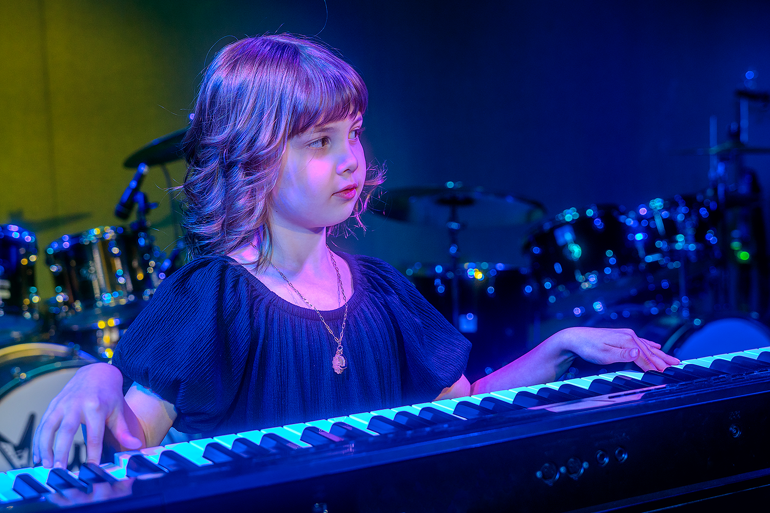 Young girl with wavy hair playing a keyboard on stage, illuminated by colorful stage lighting, with drums in the background.