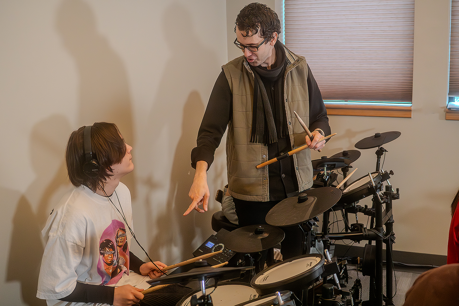 Music instructor teaching a young woman how to play electronic drums in a room with sunlight streaming through window blinds.