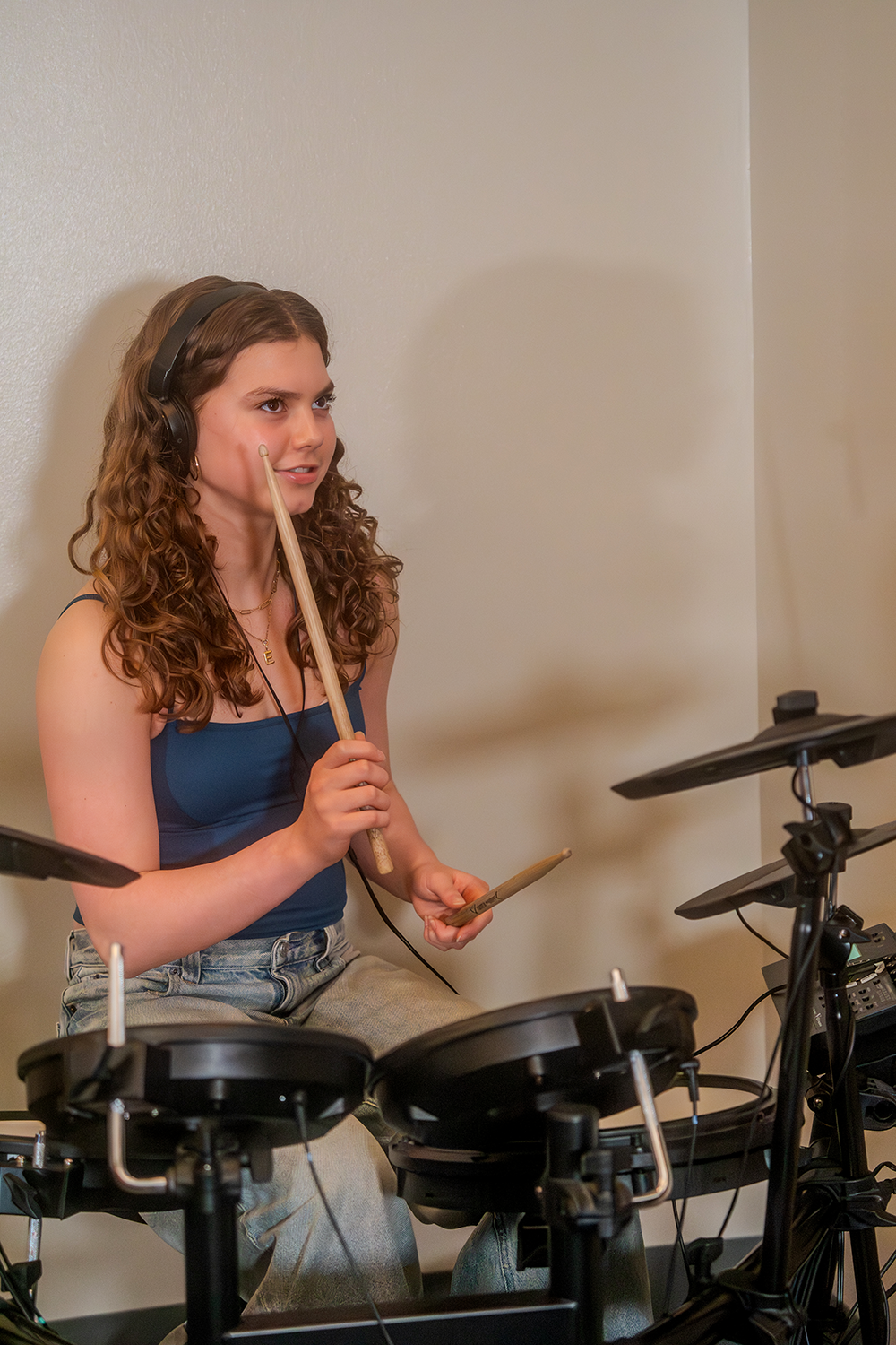 A young woman with curly hair wearing headphones plays an electronic drum set, holding drum sticks, while sitting on a stool against a beige wall.
