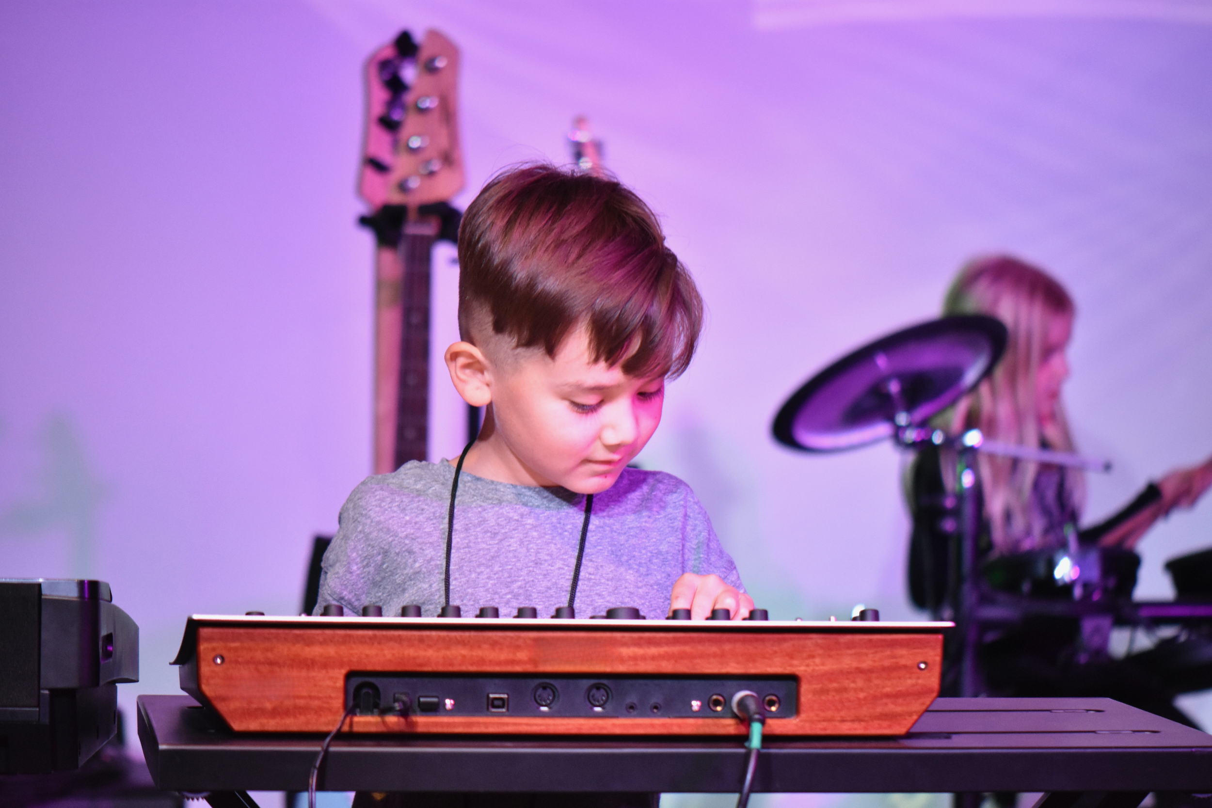 A young boy with brown hair playing a keyboard on stage, with a girl playing drums in the background and musical equipment around.