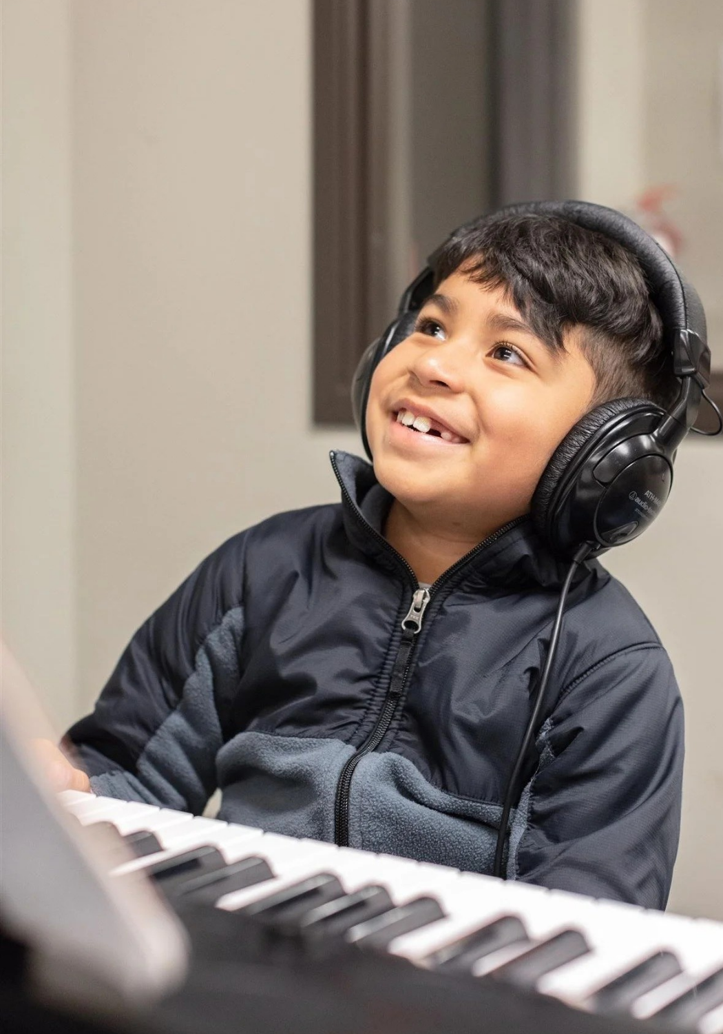 A young boy with a big smile wearing headphones, sitting at a keyboard, looking up.