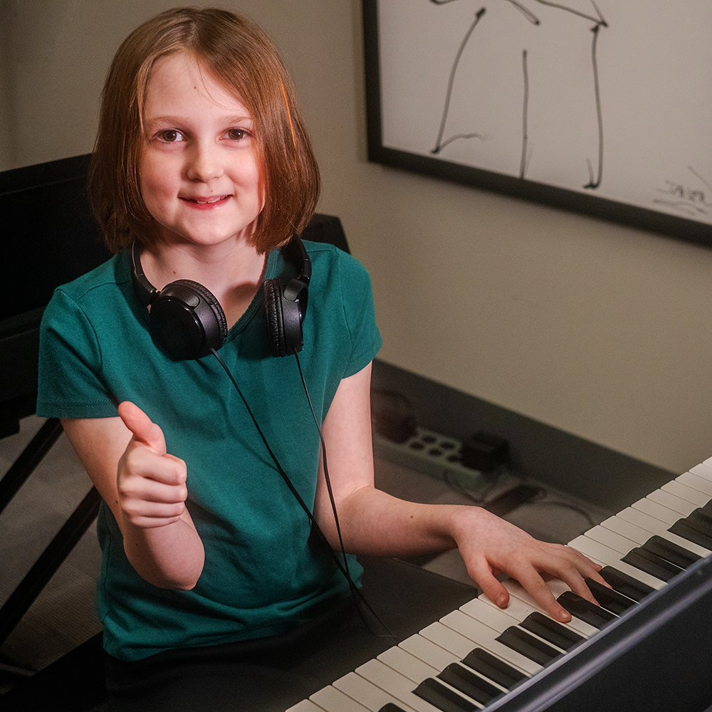 A young girl with red hair and a green shirt smiling and giving a thumbs-up while sitting at a piano with headphones around her neck, in a room with a whiteboard in the background.