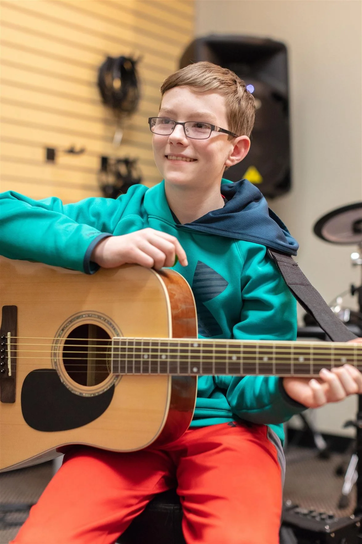 A young boy wearing glasses and a teal hoodie playing an acoustic guitar in a music room.