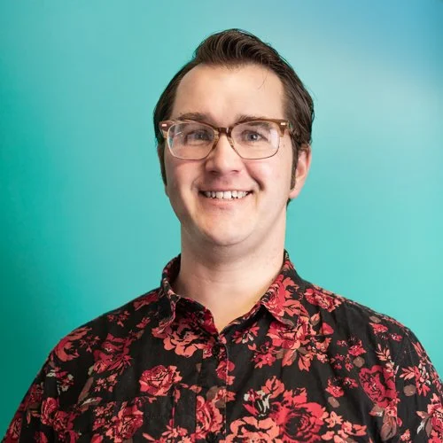 A smiling man wearing glasses and a black shirt with red floral pattern against a teal background.