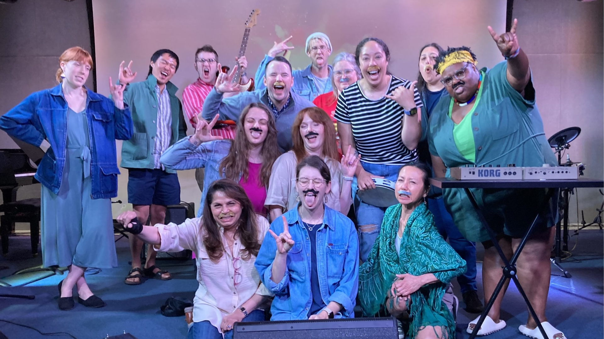 A diverse group of people on stage, some wearing fake mustaches, making hand signs, and smiling, with musical instruments and equipment around them.