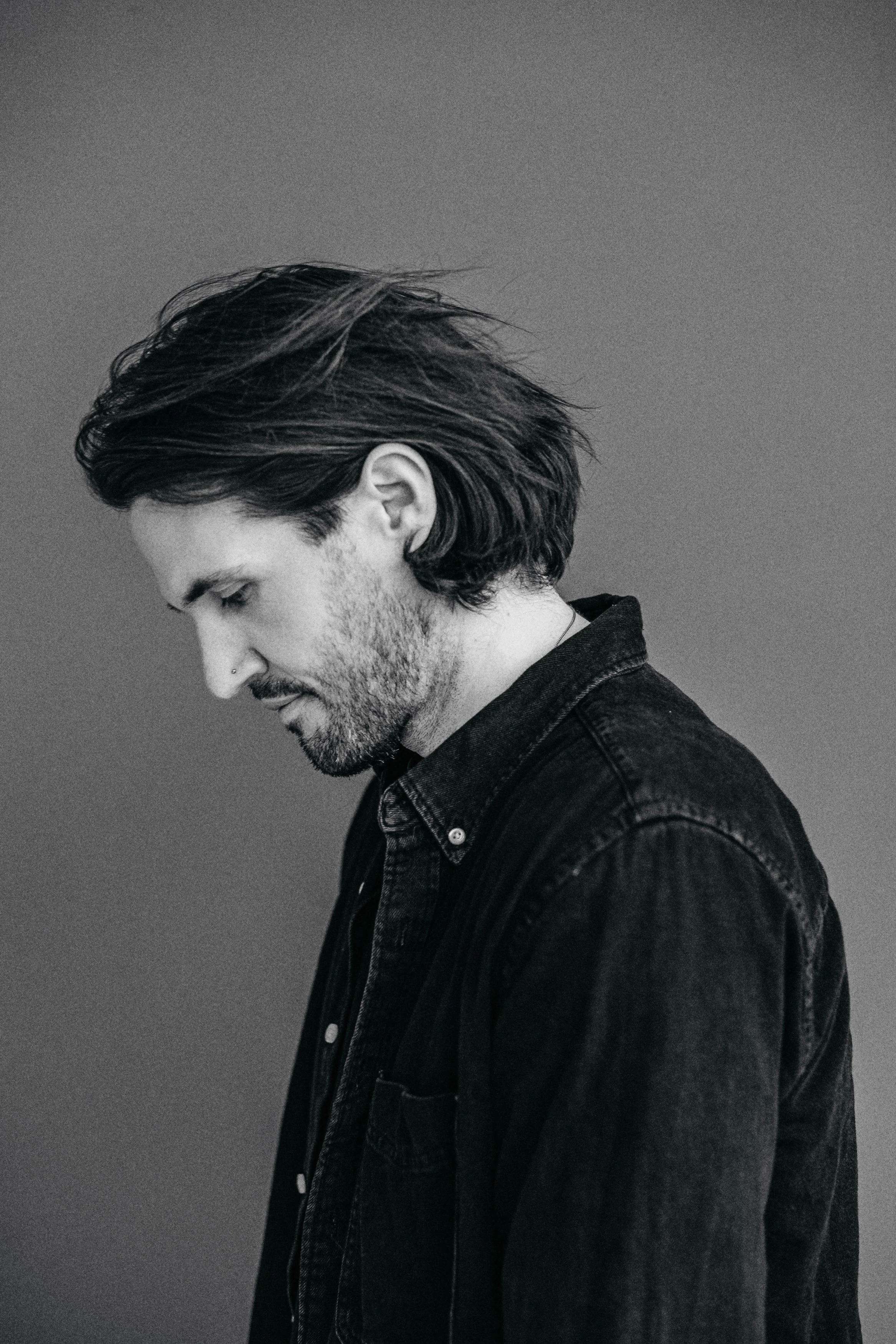 Schwarz-weiß Portrait eines Mannes mit dunklen, langen Haaren, Bart und Nasenpiercing, getragen dunkles Hemd, nach unten schauend, gegen grauen Hintergrund.