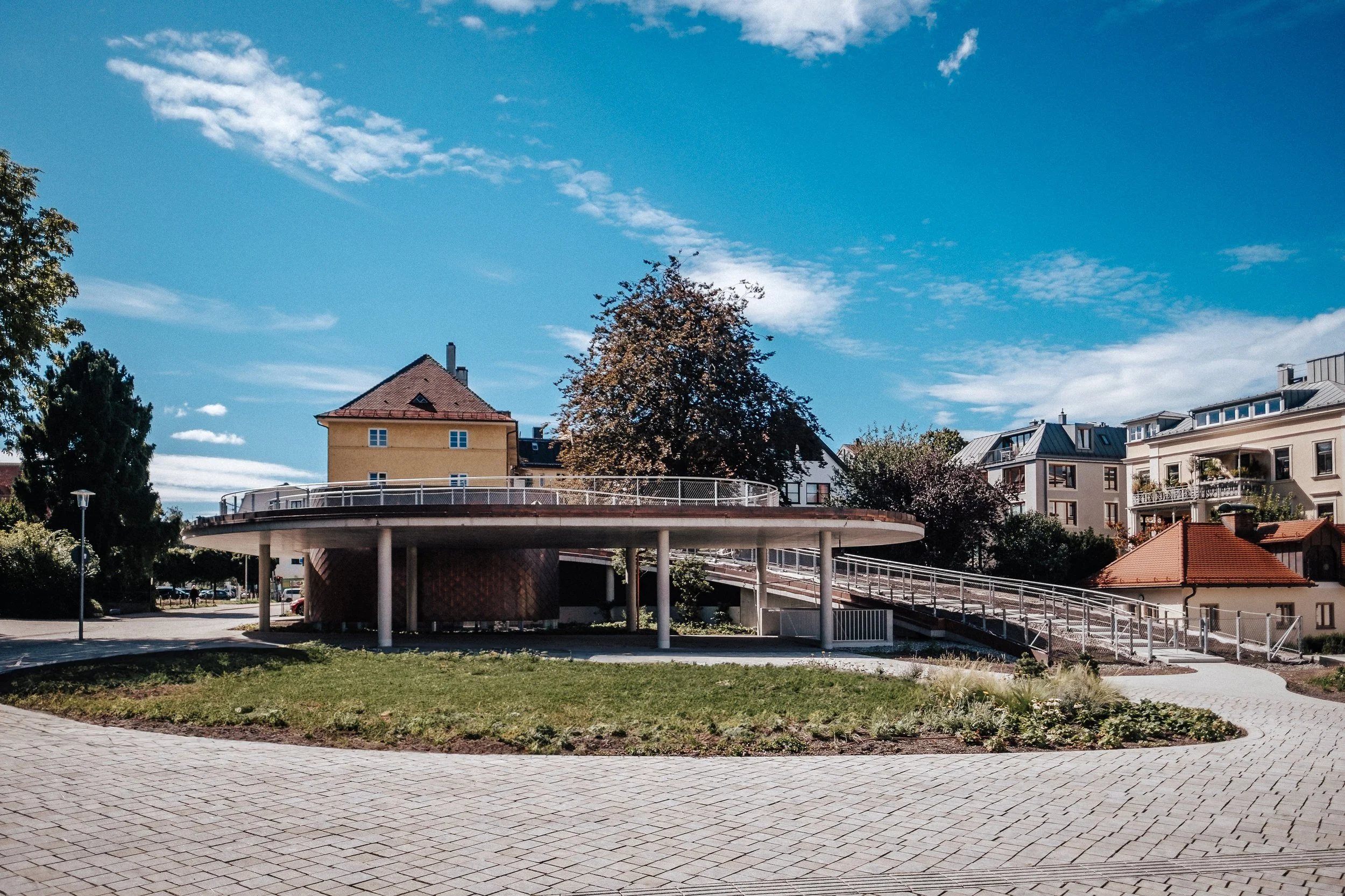 Ein moderner, runder Pavillon mit einer Rolltreppe auf der rechten Seite, umgeben von einem Park mit Wegen und Bäumen, im Hintergrund Wohnhäuser bei schönem Wetter mit blauer Himmel.