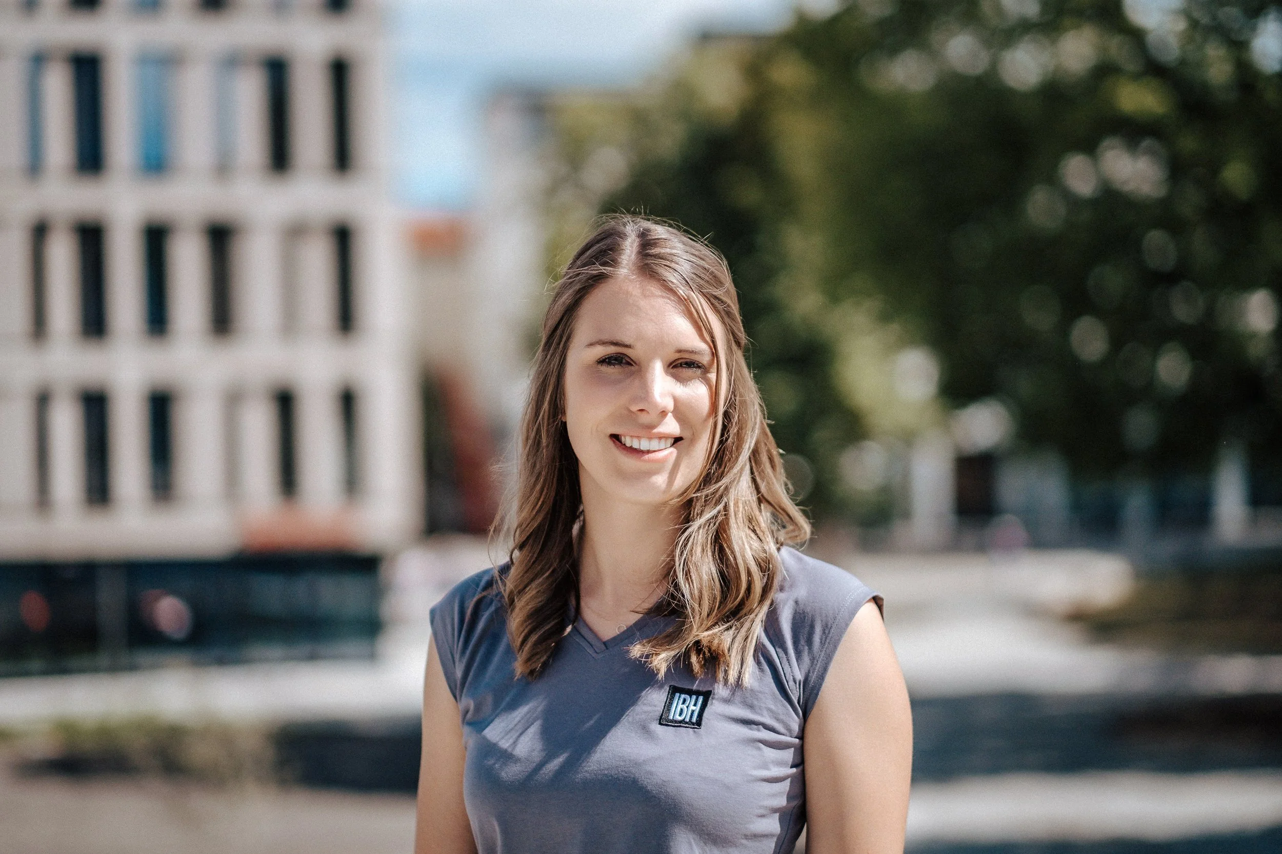 Eine junge Frau mit braunen, gewellten Haaren, trägt ein graues sportliches Oberteil mit einem Logo, steht draußen bei sonnigem Wetter vor einem Hintergrund mit Bäumen und modernen Gebäuden.