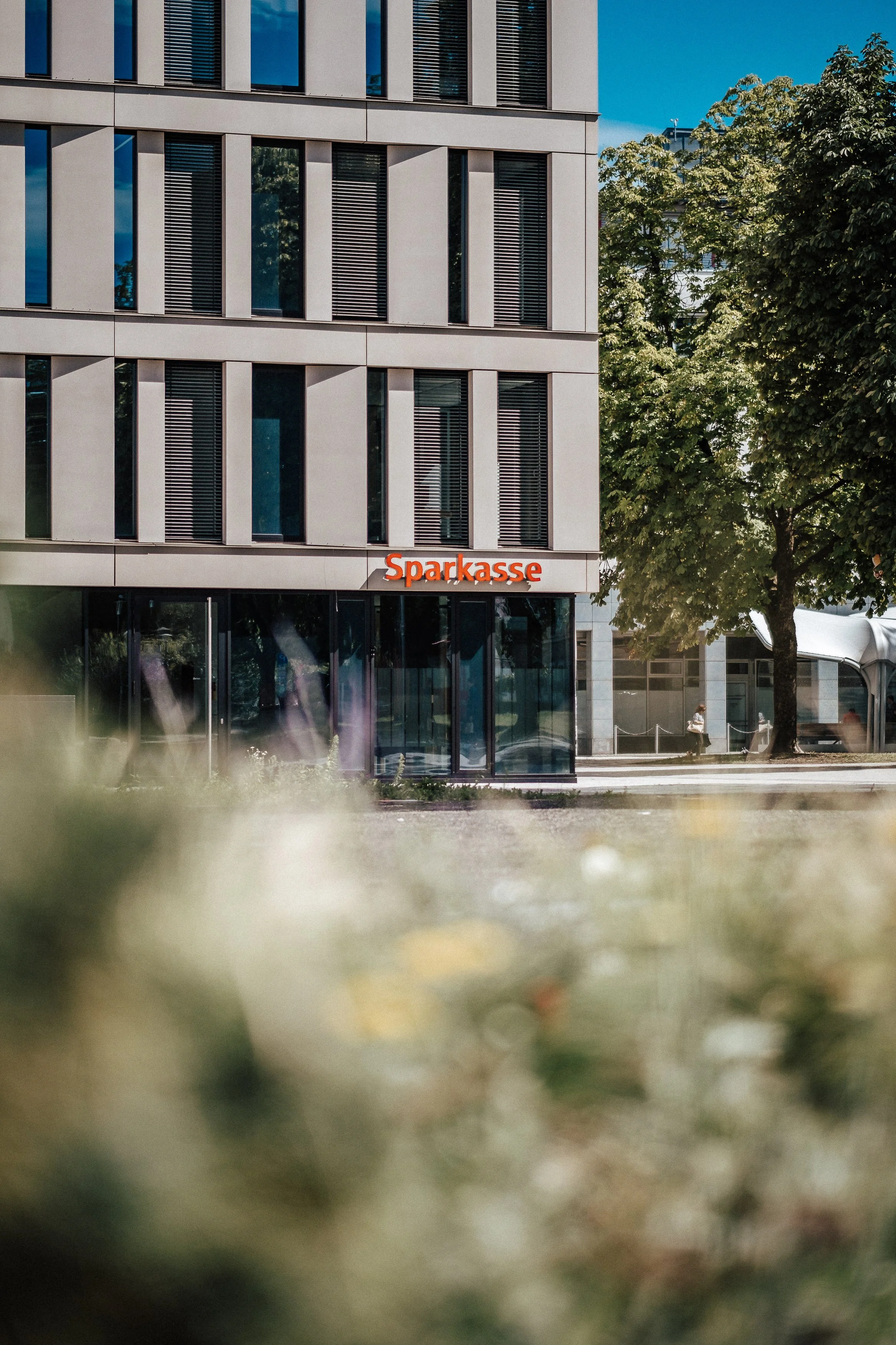 Modernes Gebäude mit einem Sparkasse-Geschäft im Erdgeschoss, umgeben von Bäumen, bei sonnigem Wetter.