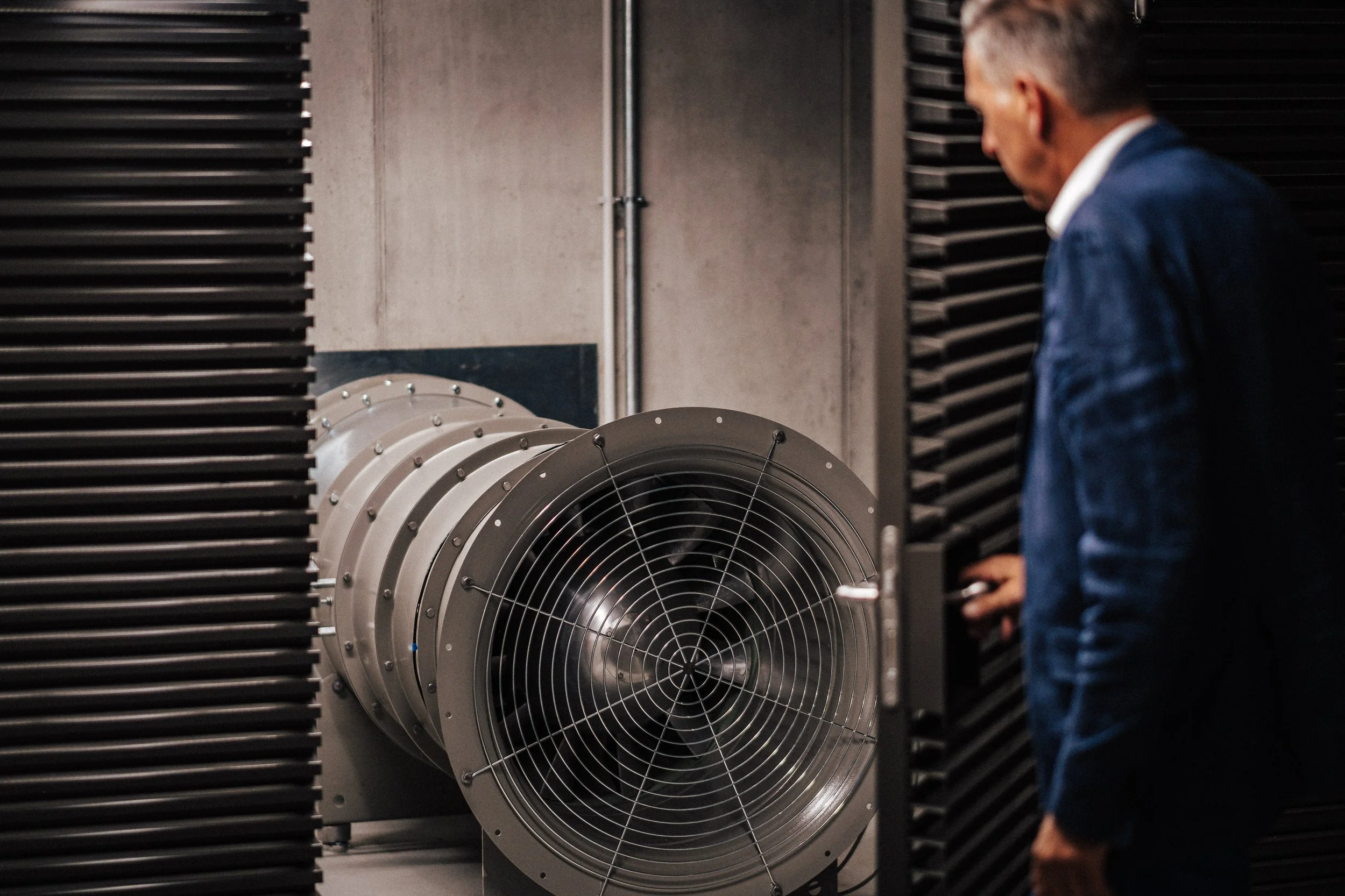 Ein Mann in dunklem Anzug mit weißem Hemd steht vor großen technischen Anlagen, die wie Luft- oder Wasserkühler aussehen, in einem technischen Raum.