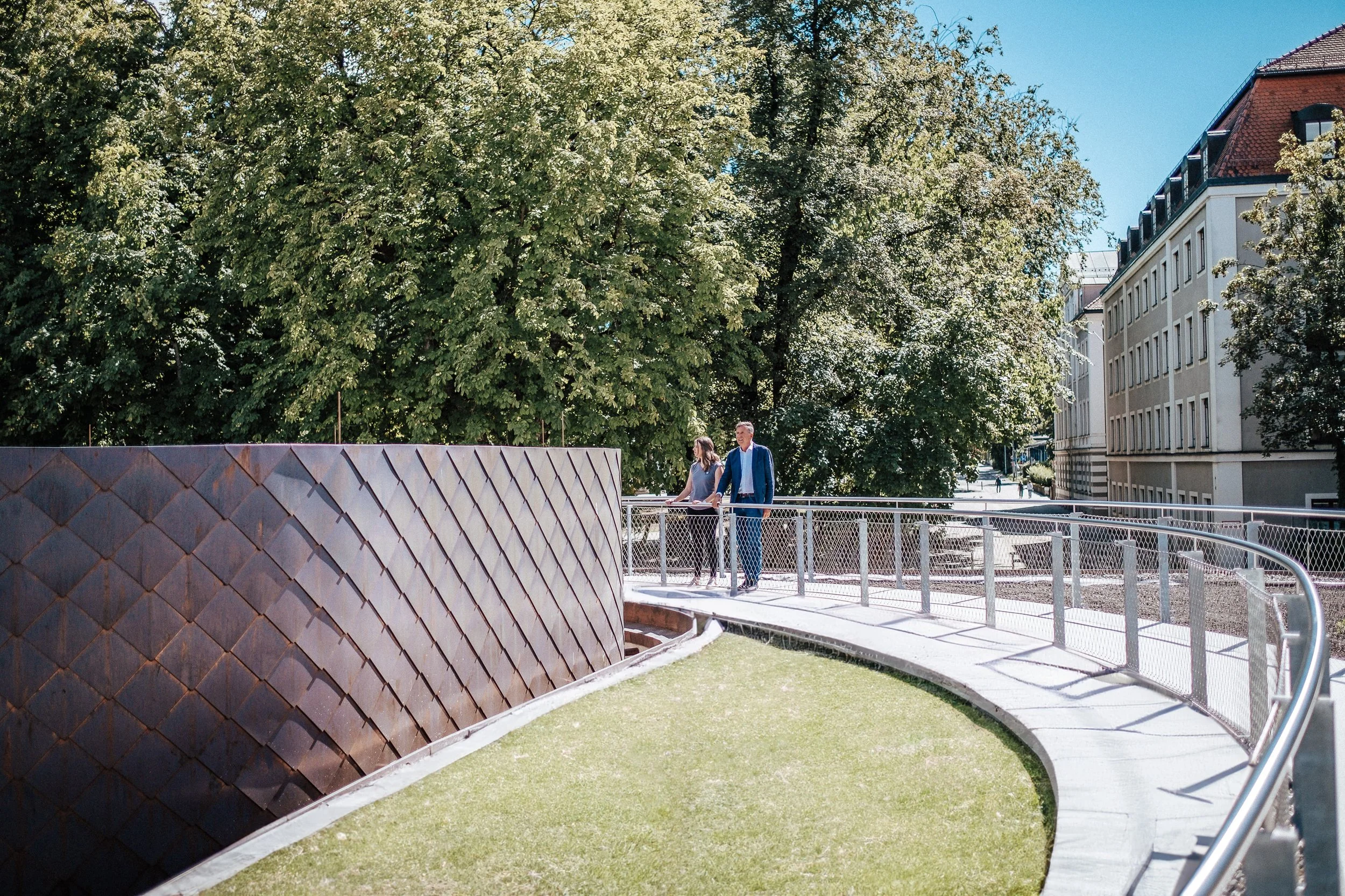 Zwei Menschen, ein Mann in einem blauen Anzug und eine Frau, gehen auf einen erhöhten, betonierten Weg in einem urbanen Park mit grüner Rasenfläche, umgeben von großen Bäumen und Gebäuden.