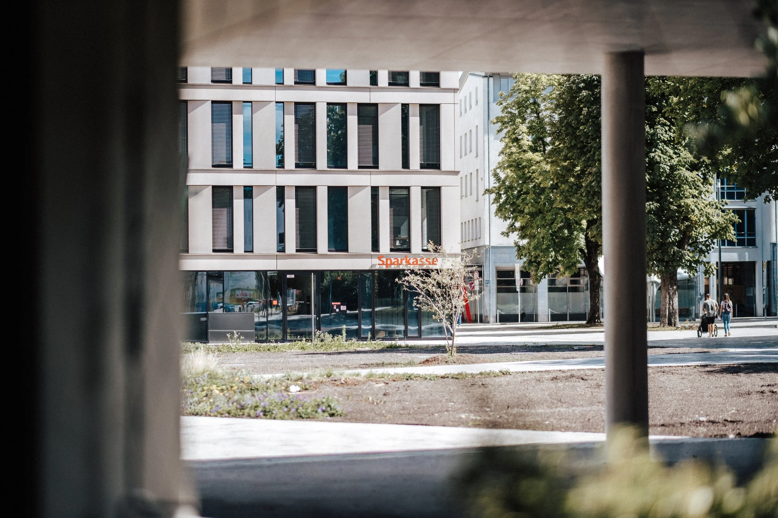 Blick auf eine moderne städtische Straßenszene durch eine Aussparung zwischen zwei Säulen. Es gibt ein mehrstöckiges Gebäude mit großen Fenstern und ein Geschäft mit dem Schild 'Sparkasse'. Es sind Bäume, eine kleine Blume und einige Fußgänger zu seh