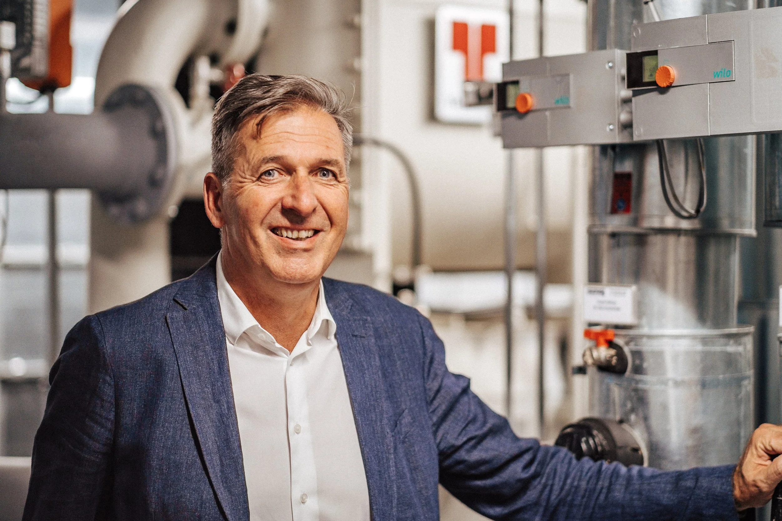 Ein lachender älterer Mann in einem blauen Anzug steht in einer technischen oder industriellen Umgebung mit Maschinen im Hintergrund.