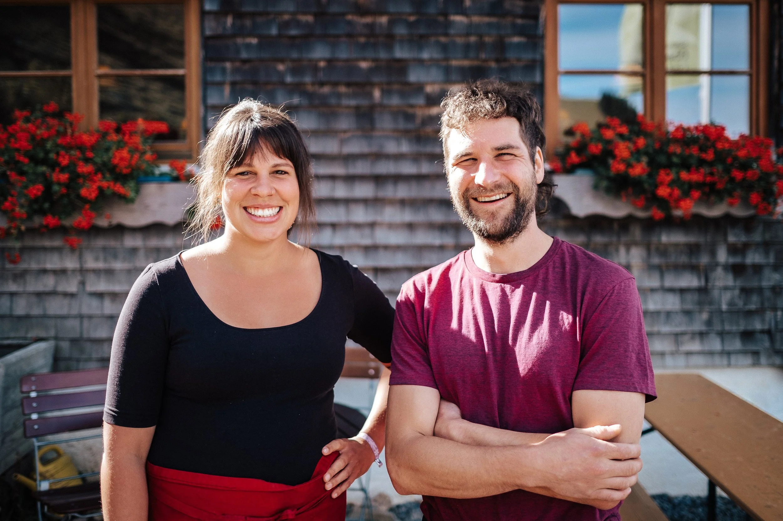 Ein Mann und eine Frau stehen nebeneinander vor einem Holzhaus mit roten Blumen vor den Fenstern, lächeln in die Kamera, sonniger Tag.