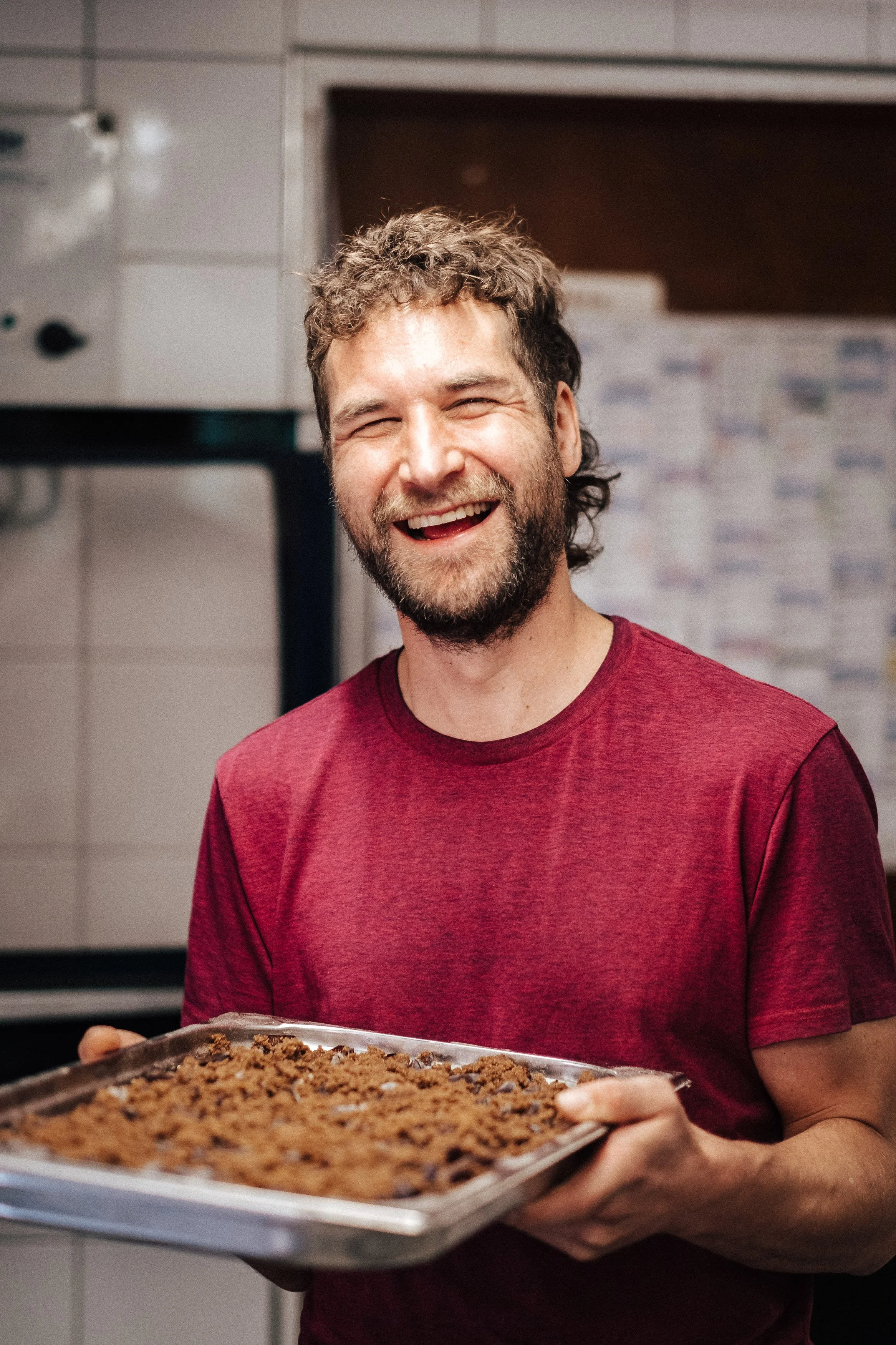 Ein lachender Mann mit Bart und dunklen, lockigen Haaren hält ein Backblech mit frisch gebackenem Keksen in einer Küche.