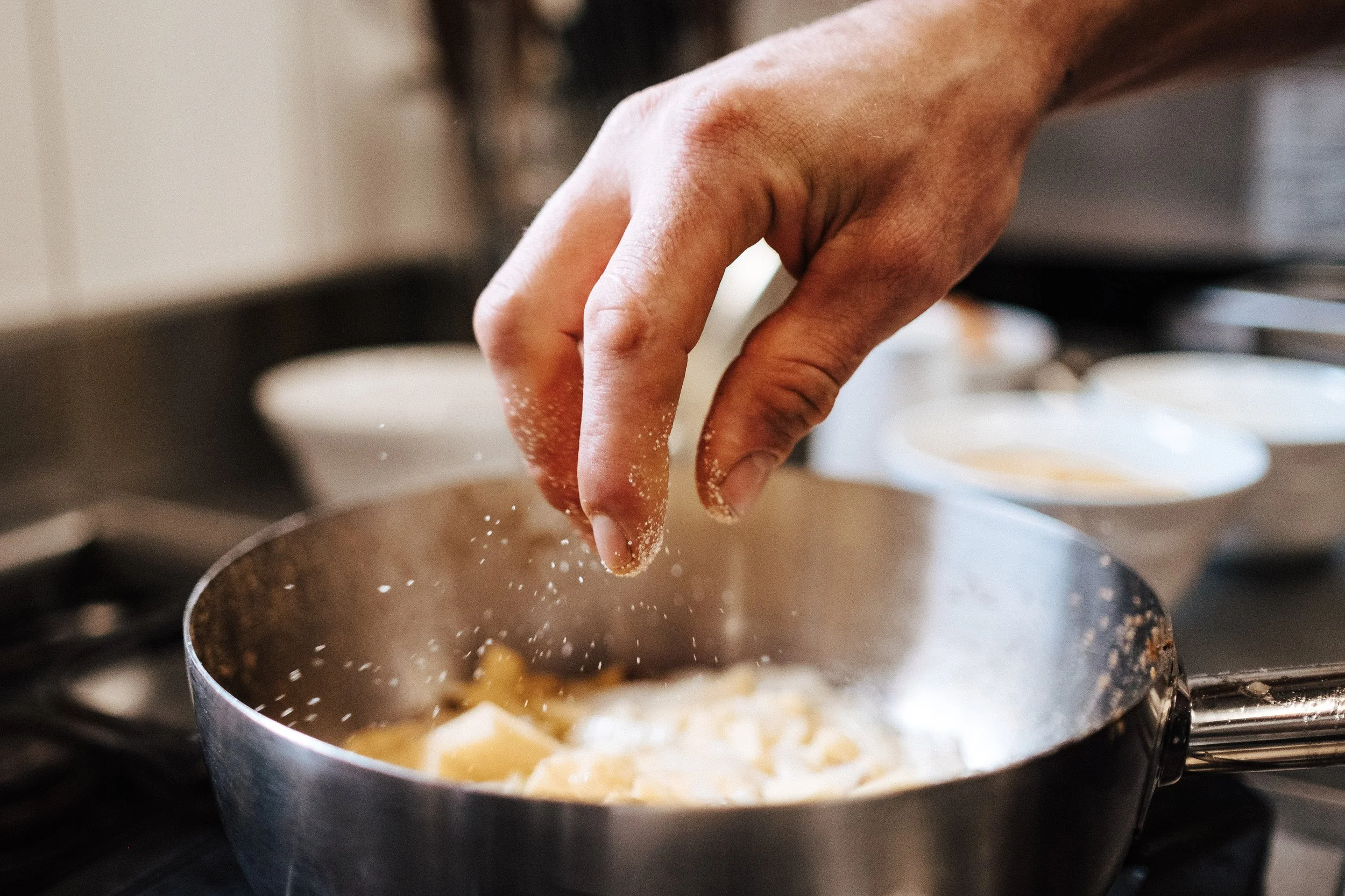 Nahaufnahme einer Hand, die Salz oder Gewürze in eine Pfanne mit gekochten Nudeln streut.